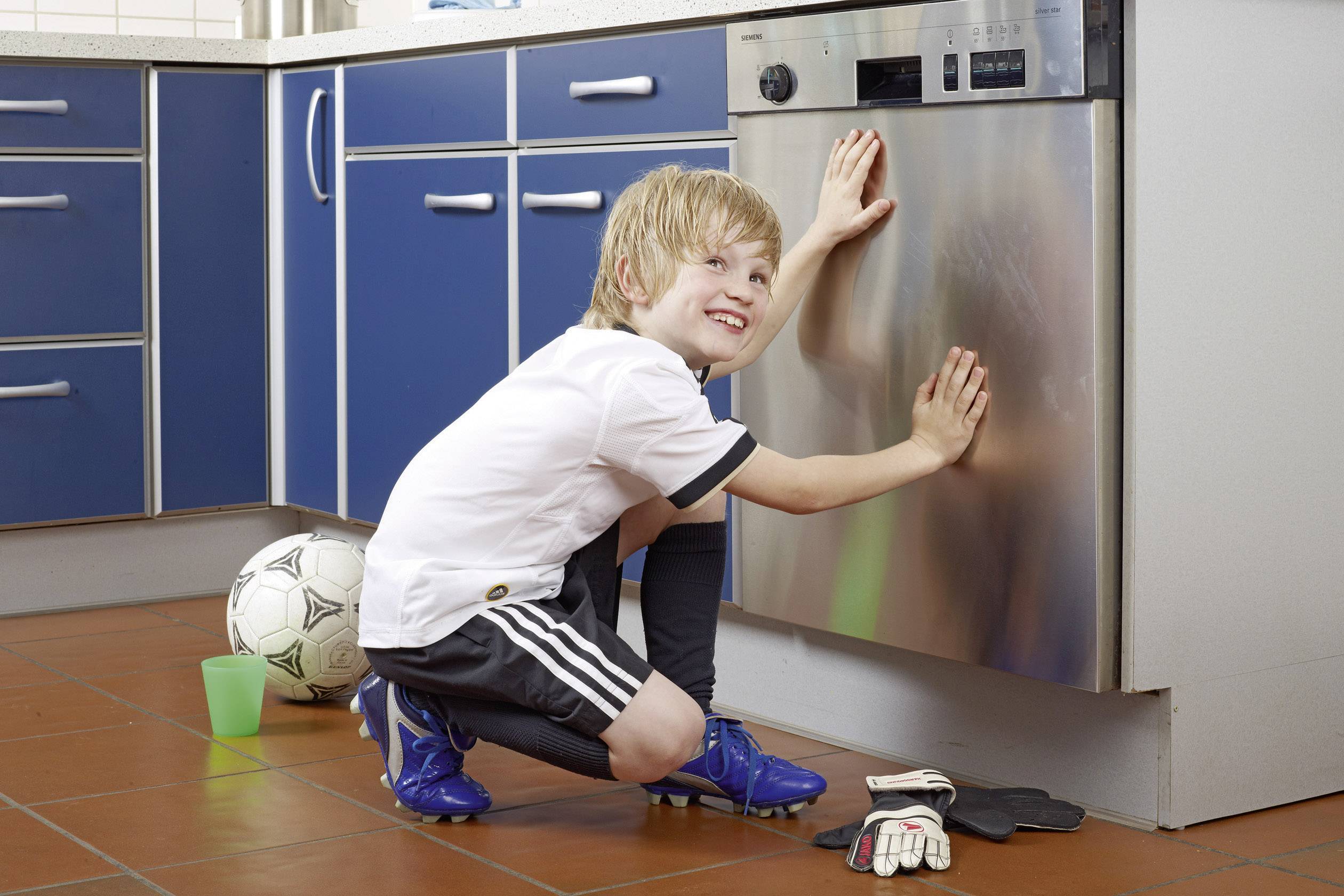 A boy in sportswear is cleaning a dishwasher in a kitchen, with a football and goalkeeper's gloves lying nearby.