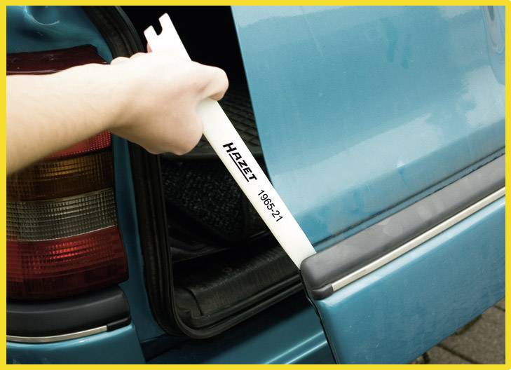 A hand is using a plastic wedge tool to remove the rear lights of a blue car. The tool bears the inscription 