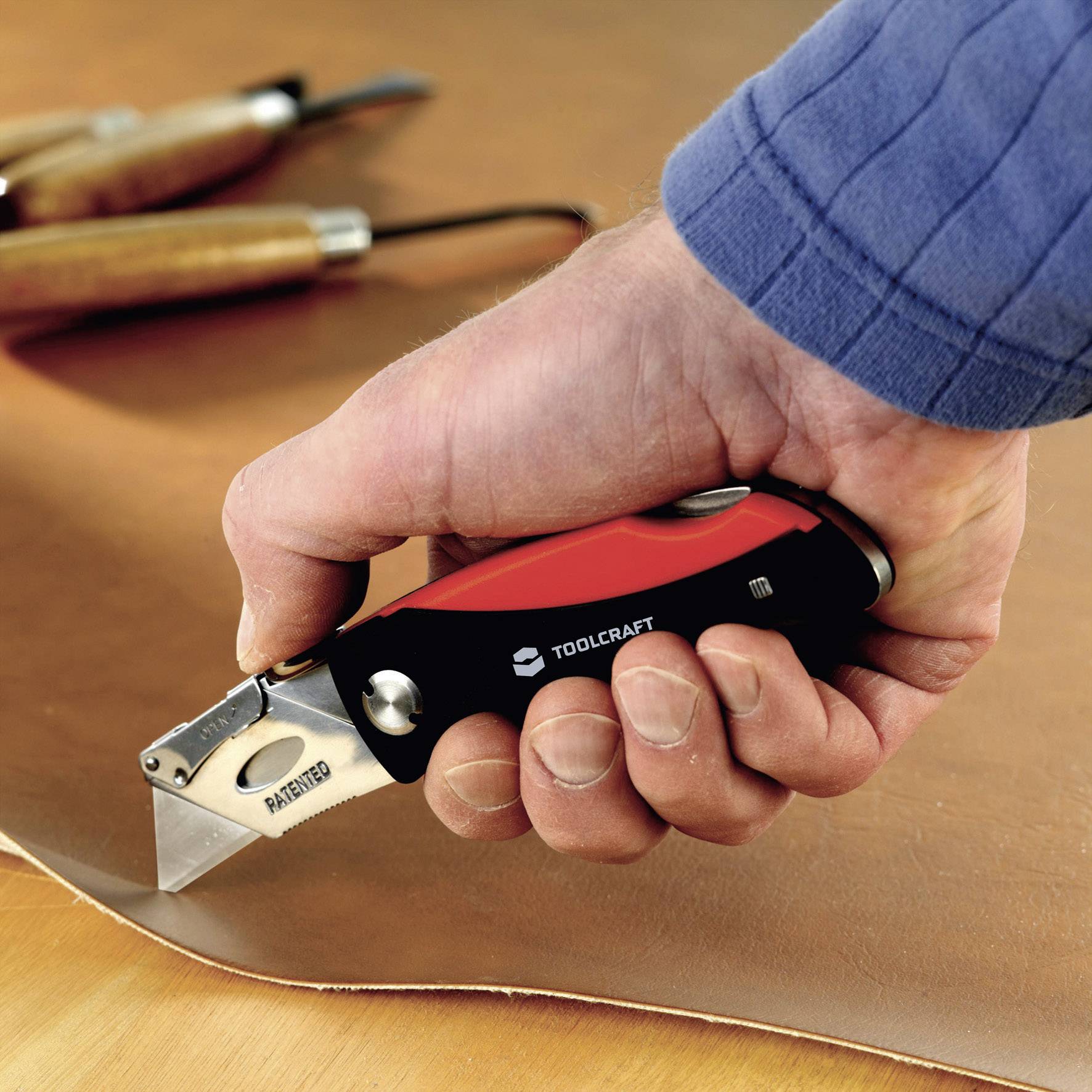 A hand holds a sharp carpet knife and cuts a piece of thick leather. Wooden tools can be seen in the background.