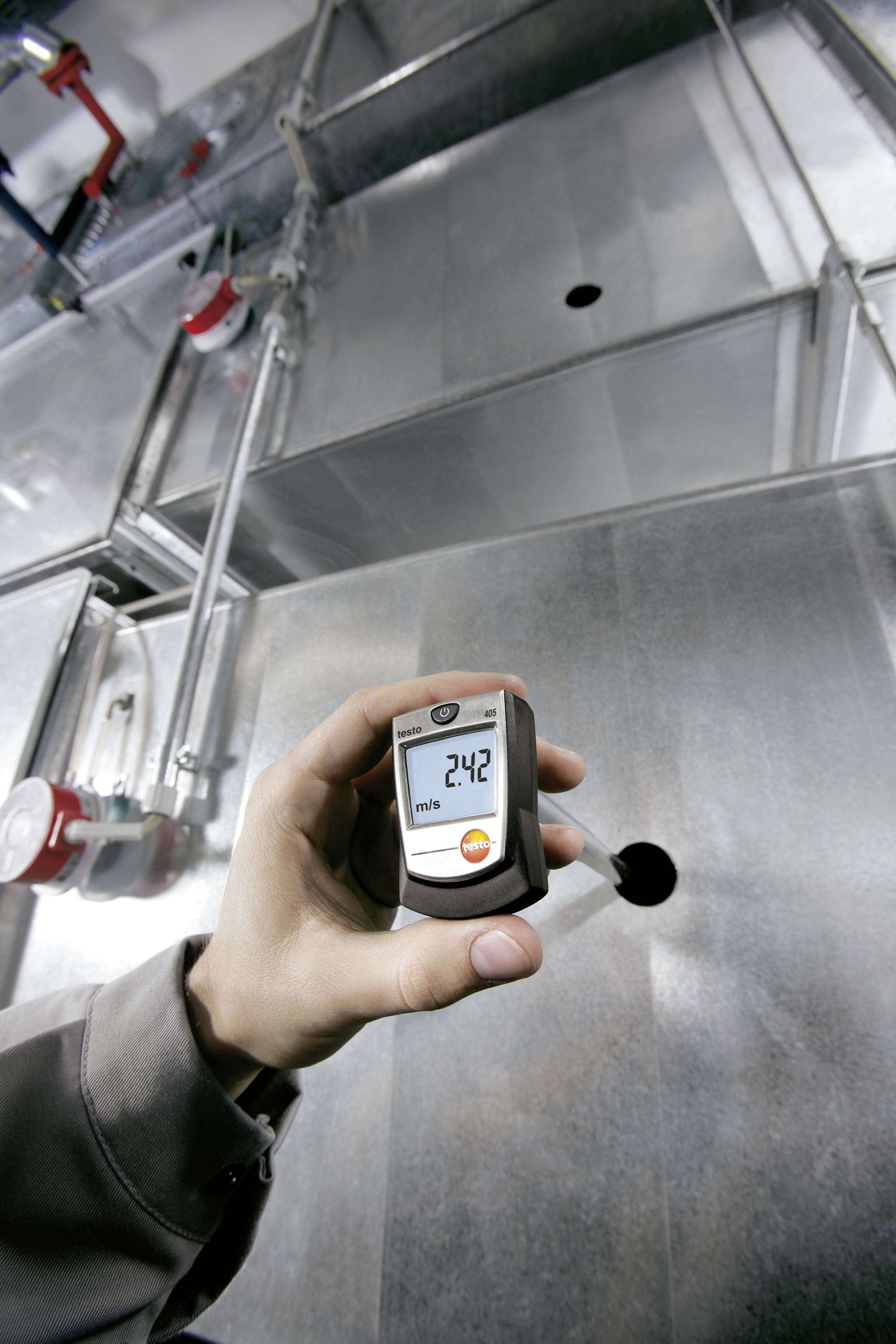 Une main tient un appareil de mesure numérique affichant '24,2'. Des conduits de ventilation industriels sont visibles en arrière-plan.