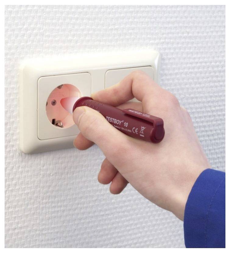 Une main tient un testeur de tension rouge contre une prise électrique. Le testeur émet une lumière rouge, indiquant la présence de tension.