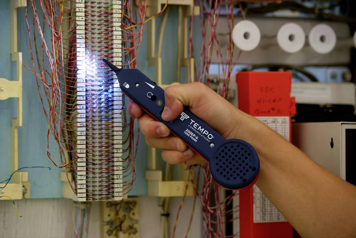 Une main tient un appareil de test de télécommunications devant un boîtier de distribution avec de nombreux câbles, éclairé pour la recherche de pannes.