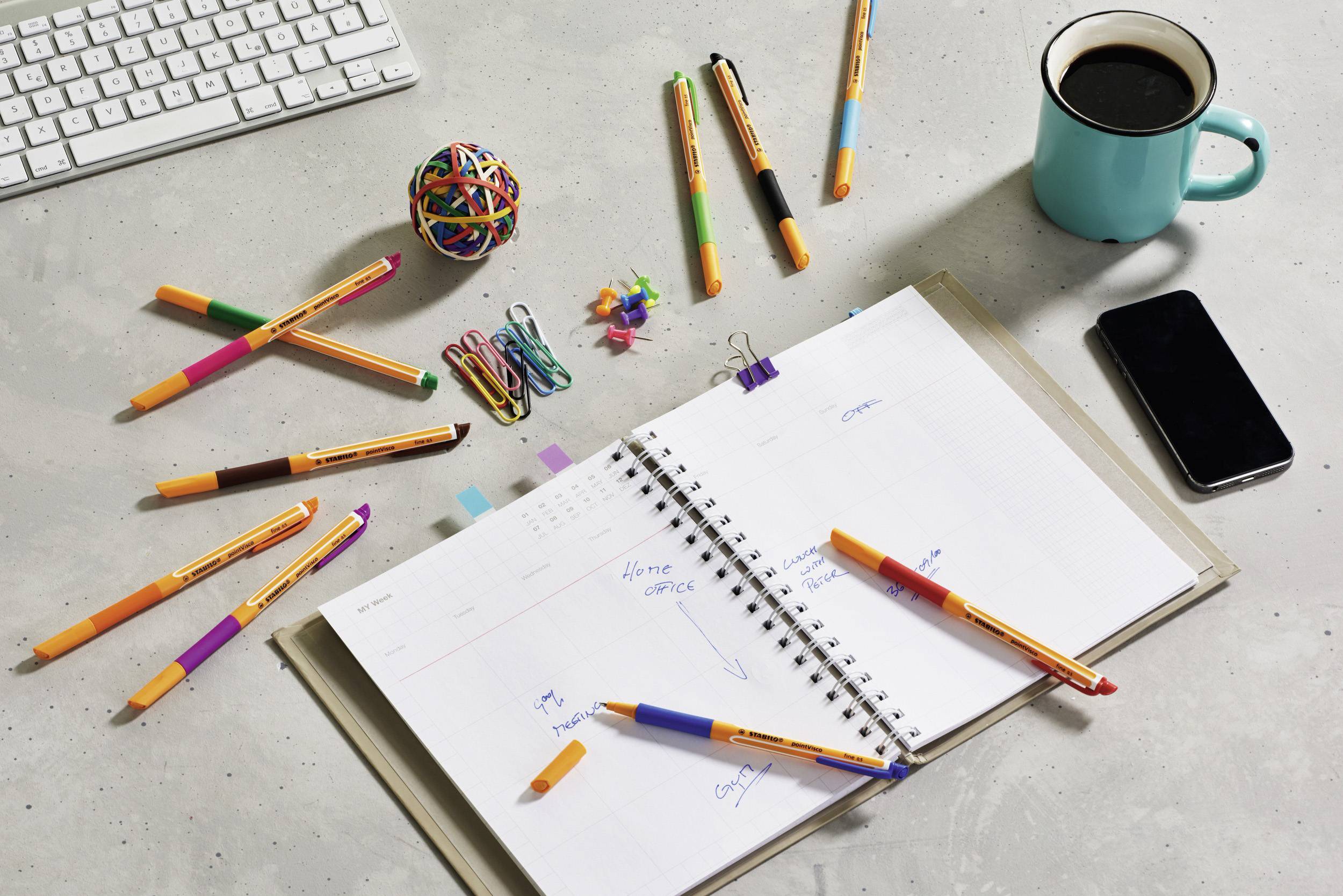 Sur un bureau, un calendrier ouvert avec des notes, divers stylos colorés et des trombones sont disposés à côté d'une tasse de café.