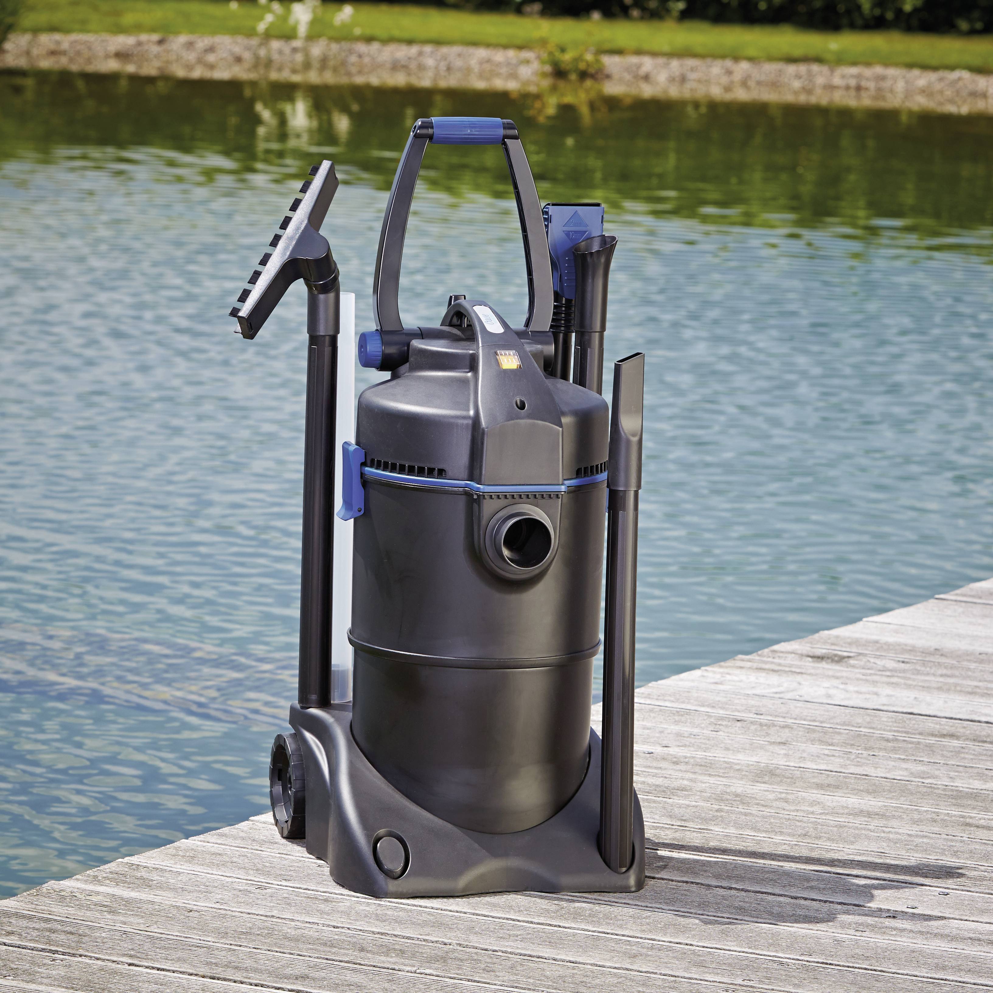 Un aspirateur noir pour bassin avec ses accessoires est posé sur une terrasse en bois à côté d'un étang. L'étang et la végétation sont visibles en arrière-plan.