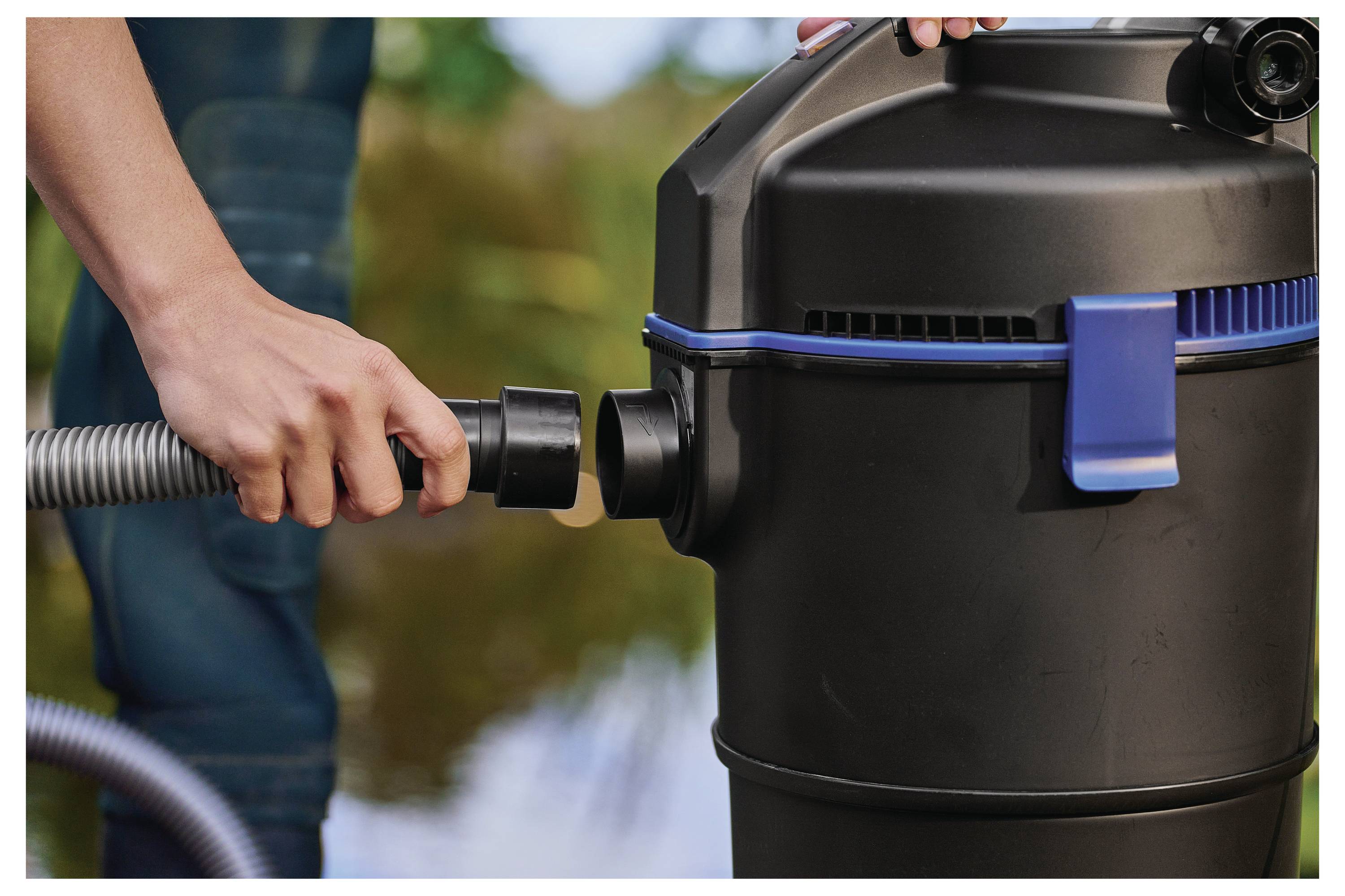 Une personne raccorde un tuyau à un aspirateur noir extérieur avec un loquet bleu, près de l'eau.