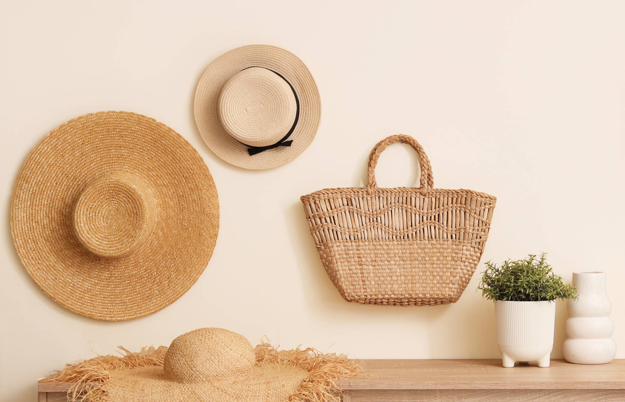 Trois chapeaux tissés et un panier en osier pendent au-dessus d'une étagère en bois contenant un chapeau de paille, une plante en pot et un vase blanc contre un mur beige.