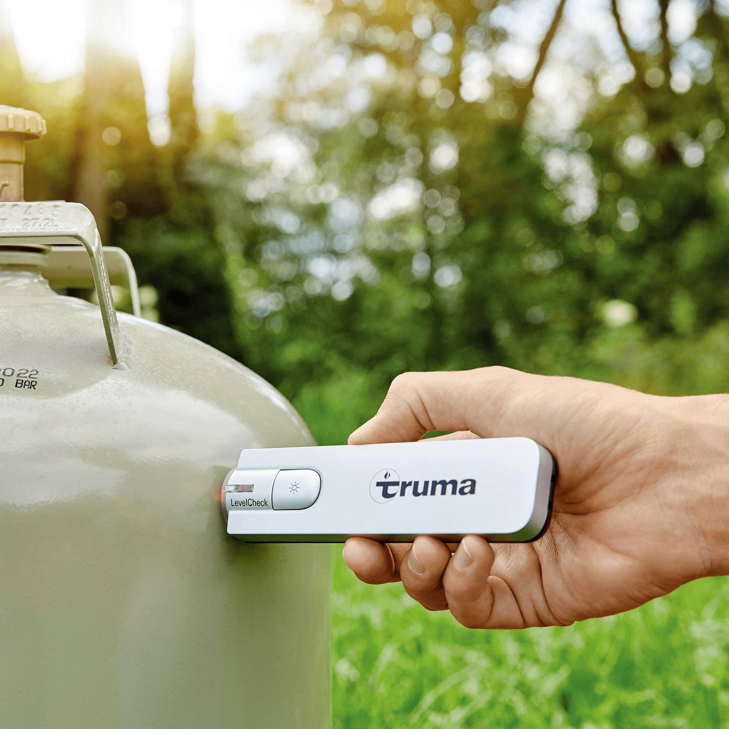 Une main tient le Truma LevelCheck sur une bouteille de gaz grise pour vérifier le niveau de remplissage, avec des arbres flous en arrière-plan.