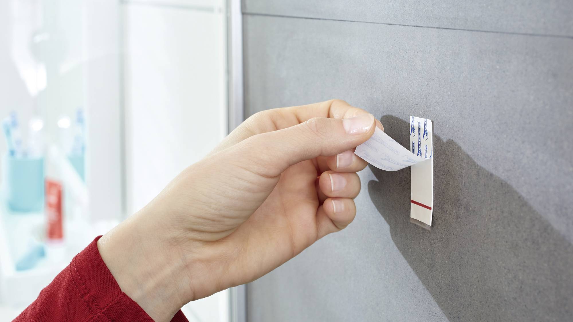Une main colle une bande de fixation sur un mur gris. Des articles de toilette sont visibles de manière floue en arrière-plan.