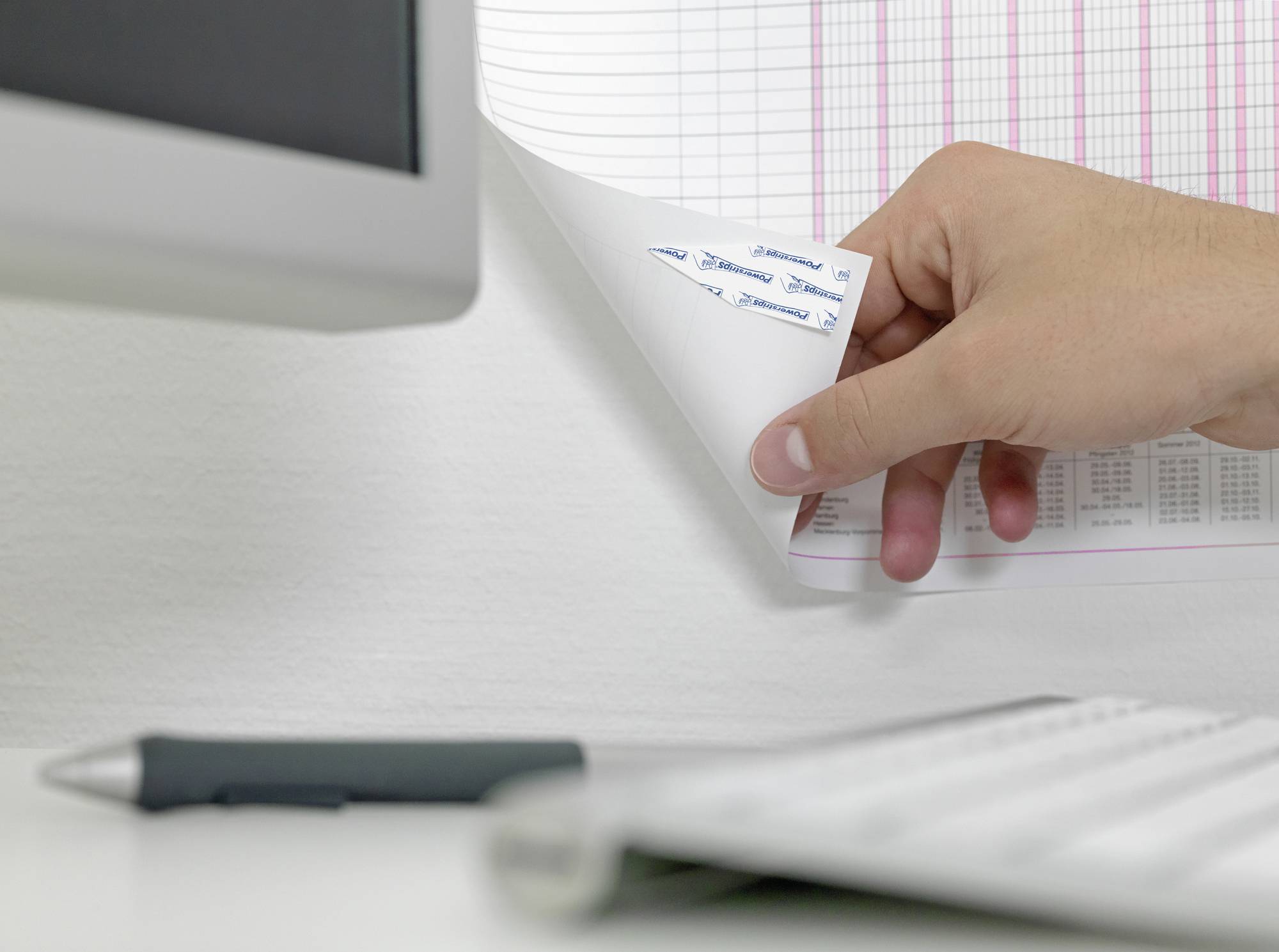 Une main feuillette une grande page de calendrier accrochée au mur. À côté, un écran flou et un stylo à bille sur un bureau.