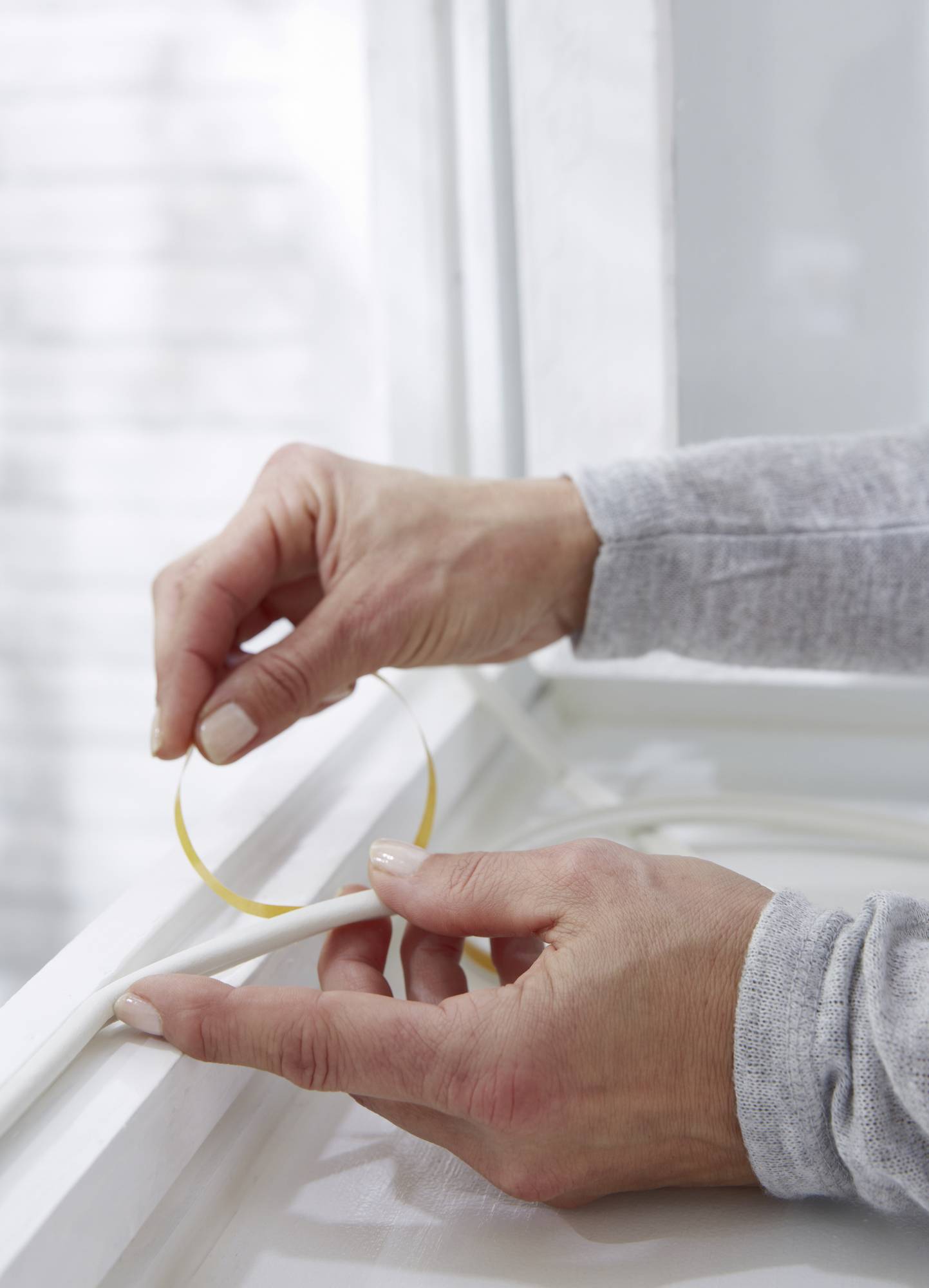 Une personne applique une fine bande jaune de joint d'étanchéité au bord d'une fenêtre blanche, probablement pour la sceller.