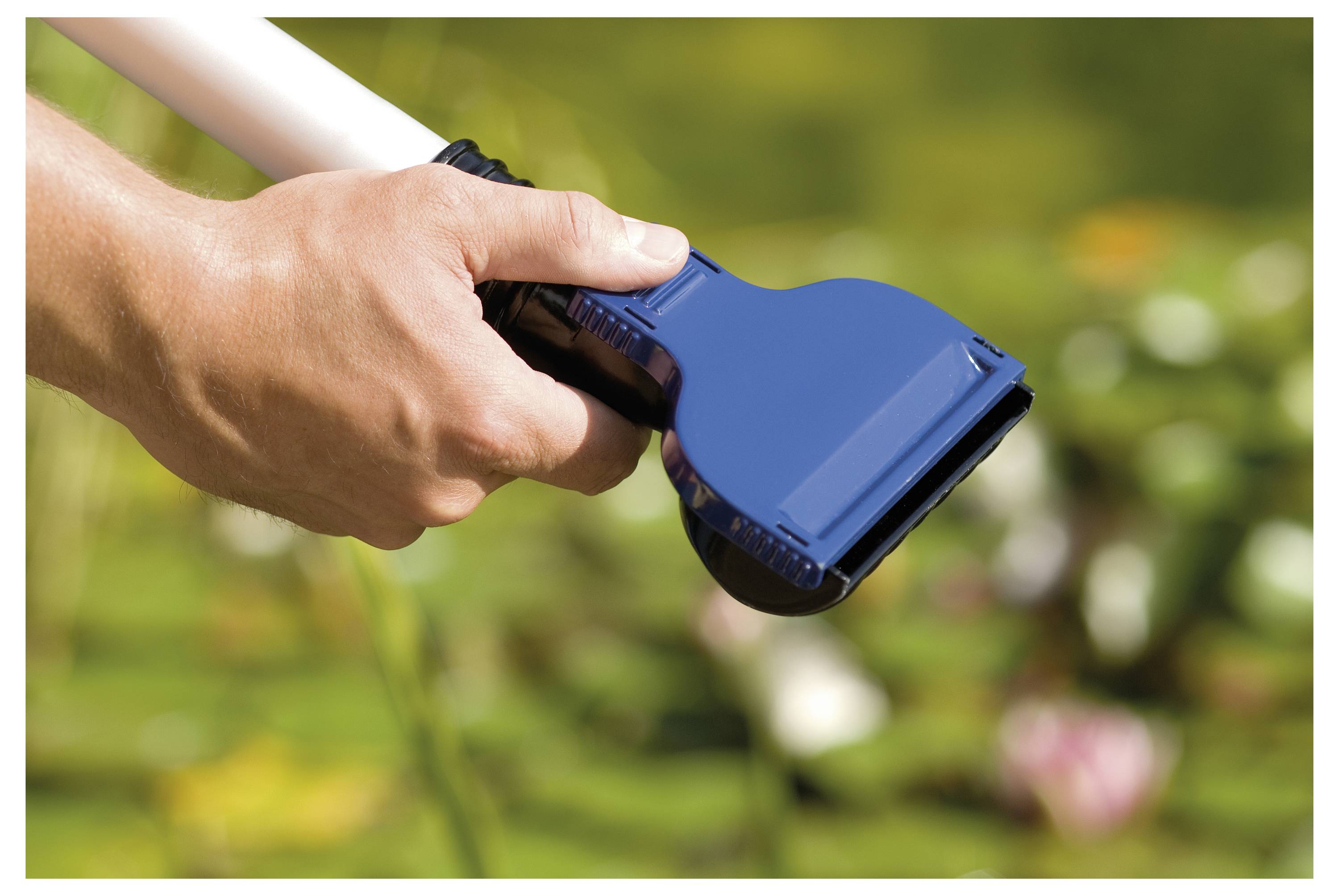 Une main tient un filet de nettoyage bleu pour piscine au-dessus d'un arrière-plan flou vert et fleuri, suggérant un nettoyage ou une maintenance extérieure.