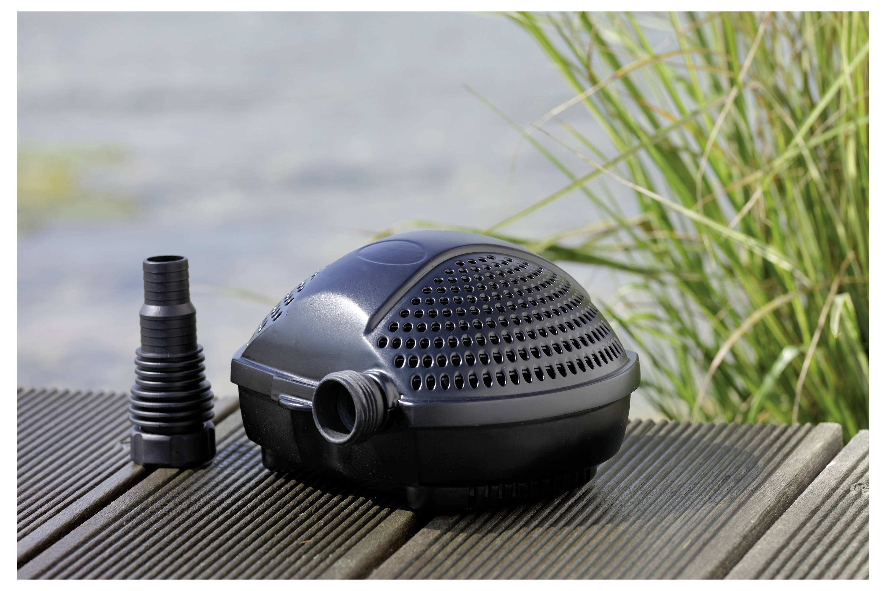 Une pompe à eau noire, conçue pour une utilisation dans les étangs, est placée sur une terrasse en bois à côté d'un plan d'eau, accompagnée d'un adaptateur de tuyau noir. De l'herbe est visible en arrière-plan.