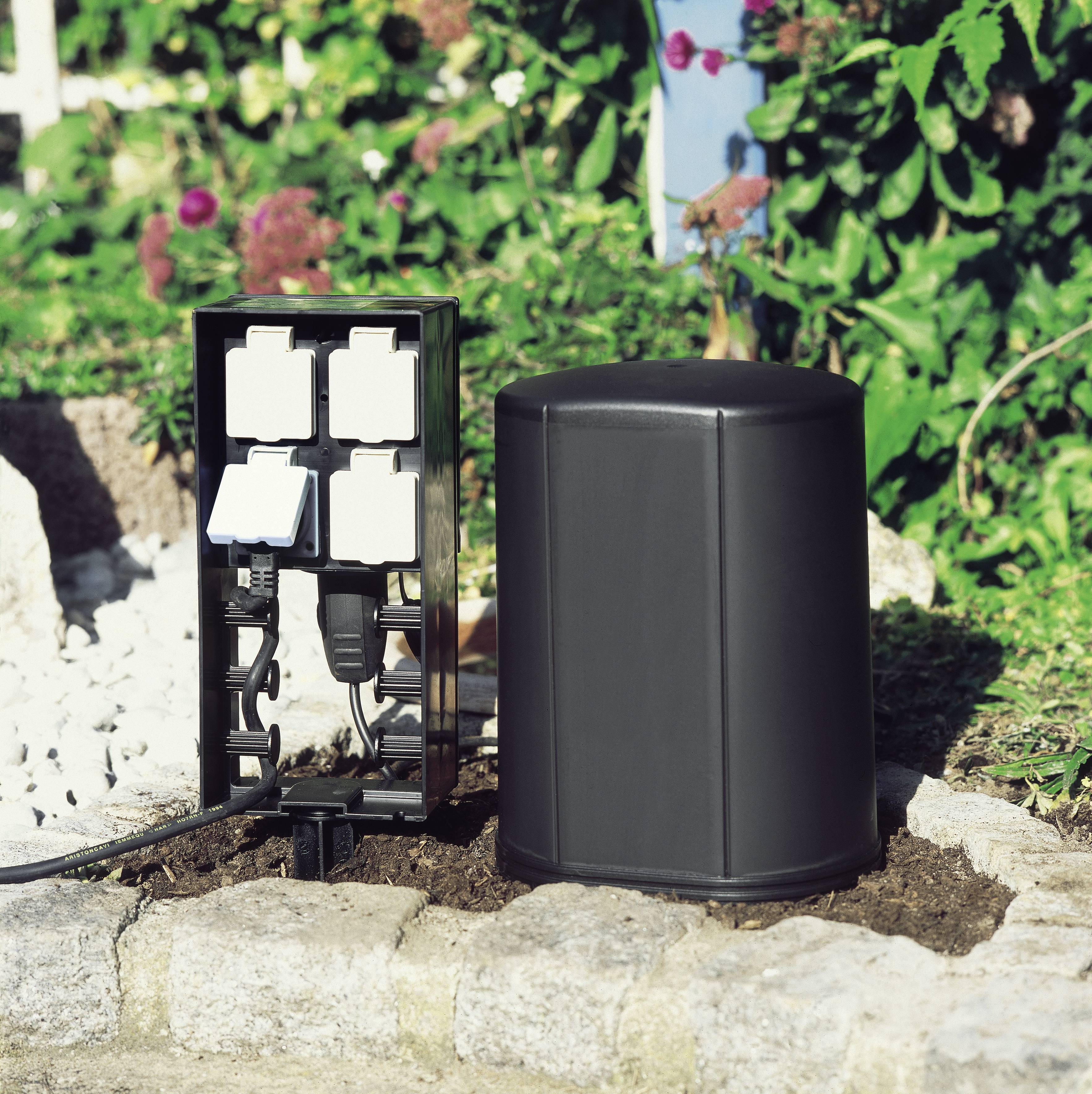 Une prise électrique extérieure avec plusieurs prises et câbles est installée dans un jardin, entourée de verdure luxuriante et de rochers.