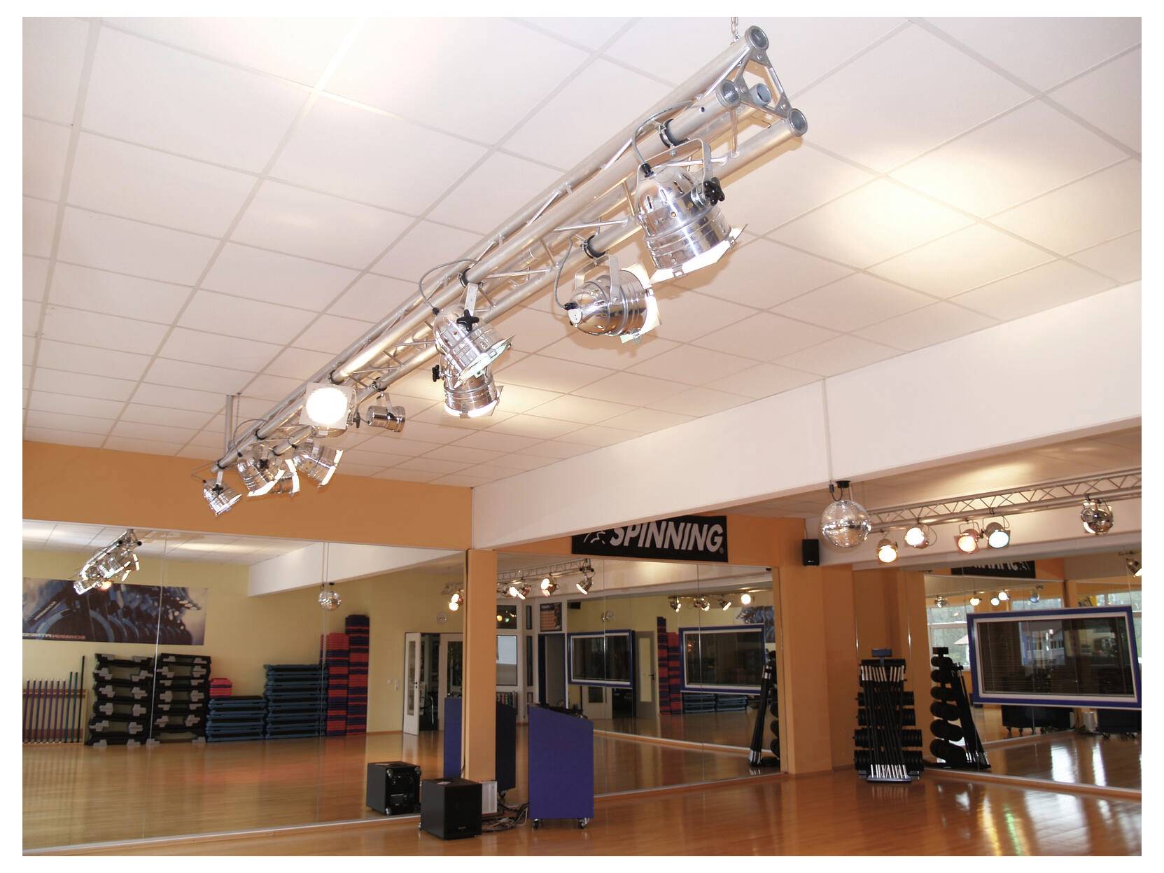 Studio de gymnase vide avec mur miroir, sol en bois, tapis d'exercice et système d'éclairage suspendu sous une enseigne « SPINNING ».