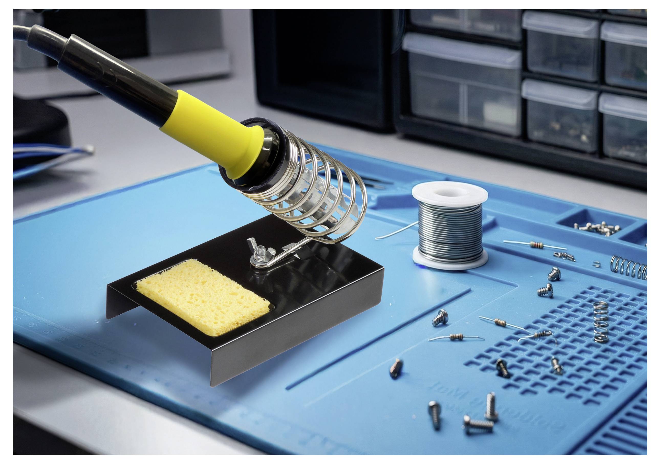 Un fer à souder avec une poignée jaune est posé dans un support sur un tapis bleu. À proximité se trouvent une bobine de soudure et des composants électroniques éparpillés.