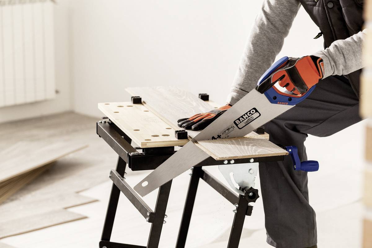 Eine Person sägt ein Holzbrett auf einer Werkbank. Sie trägt Arbeitshandschuhe und benutzt eine Handsäge.