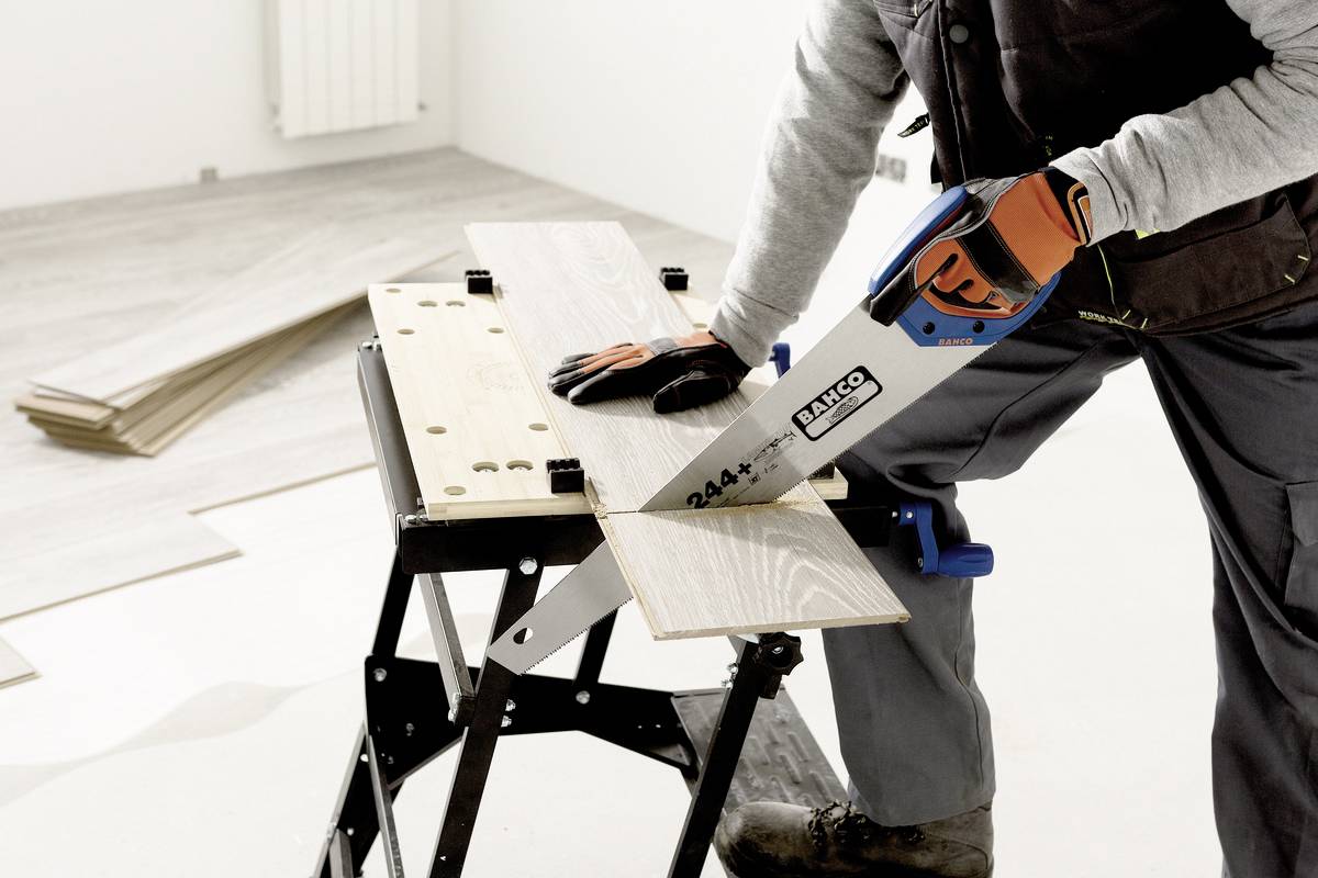 Eine Person sägt ein Holzbrett auf einem Arbeitstisch mit einer Handsäge in einem Raum. Am Boden liegen weitere Holzplatten.