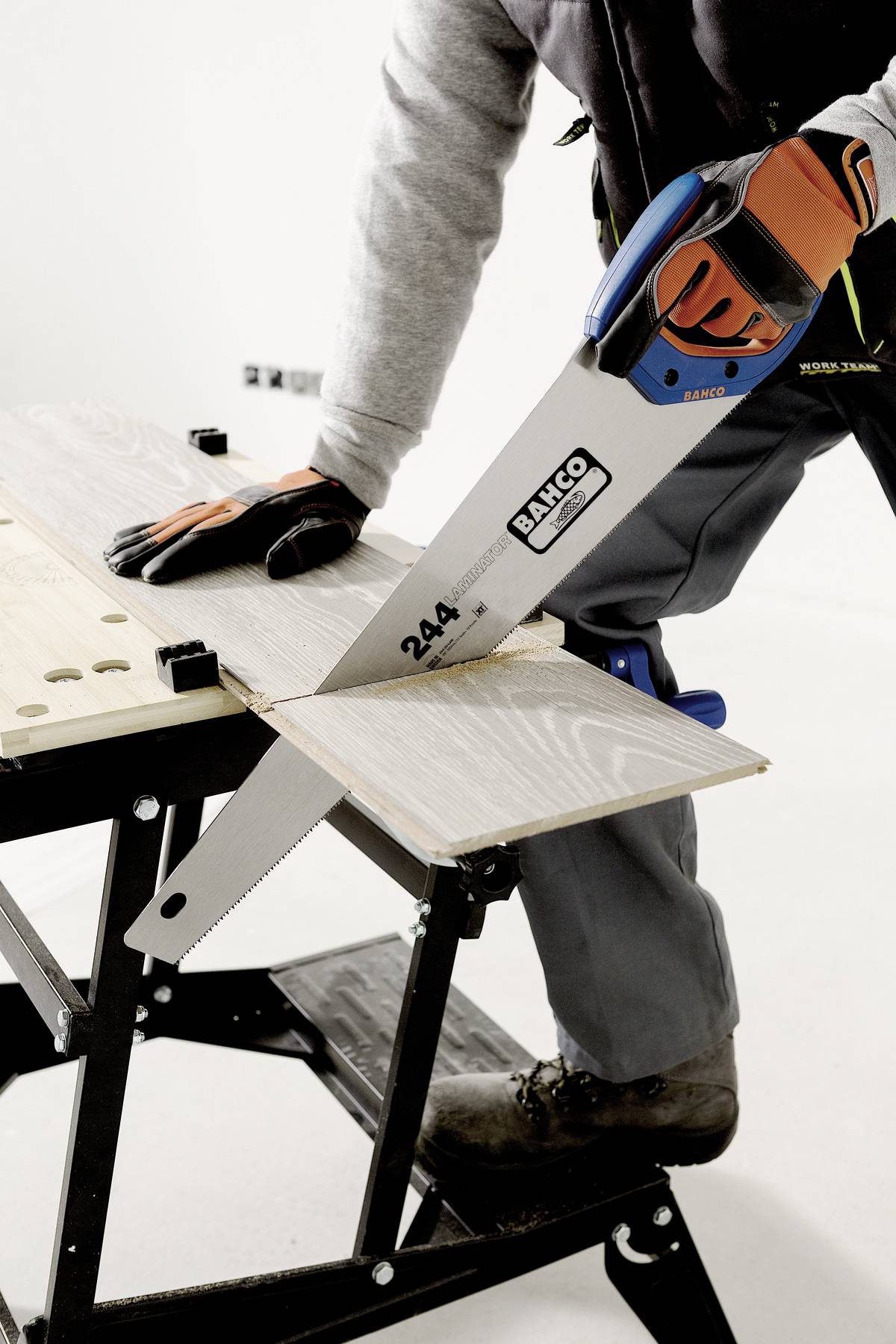Eine Person sägt ein Holzbrett auf einem Sägebock mit einer Handsäge. Die Person trägt Arbeitshandschuhe und eine Arbeitsjacke.