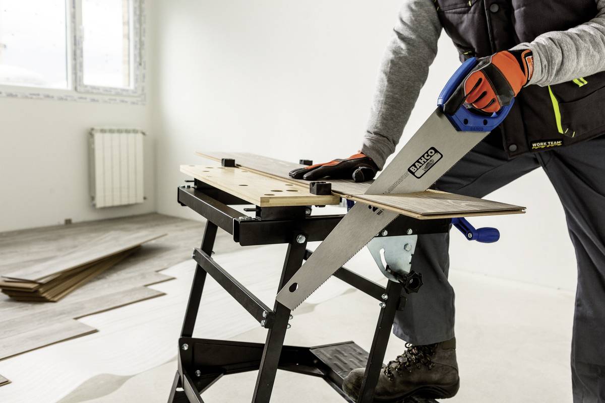 Eine Person sägt in einem leeren Raum ein Brett auf einem tragbaren Arbeitstisch. Im Hintergrund liegen Holzbodenplatten.