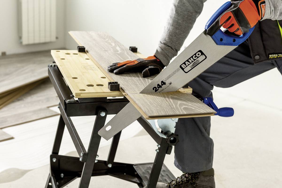 Eine Person sägt mit einer Handsäge ein Holzbrett auf einer Arbeitsbank. Sicherheitskleidung und Handschuhe werden getragen.
