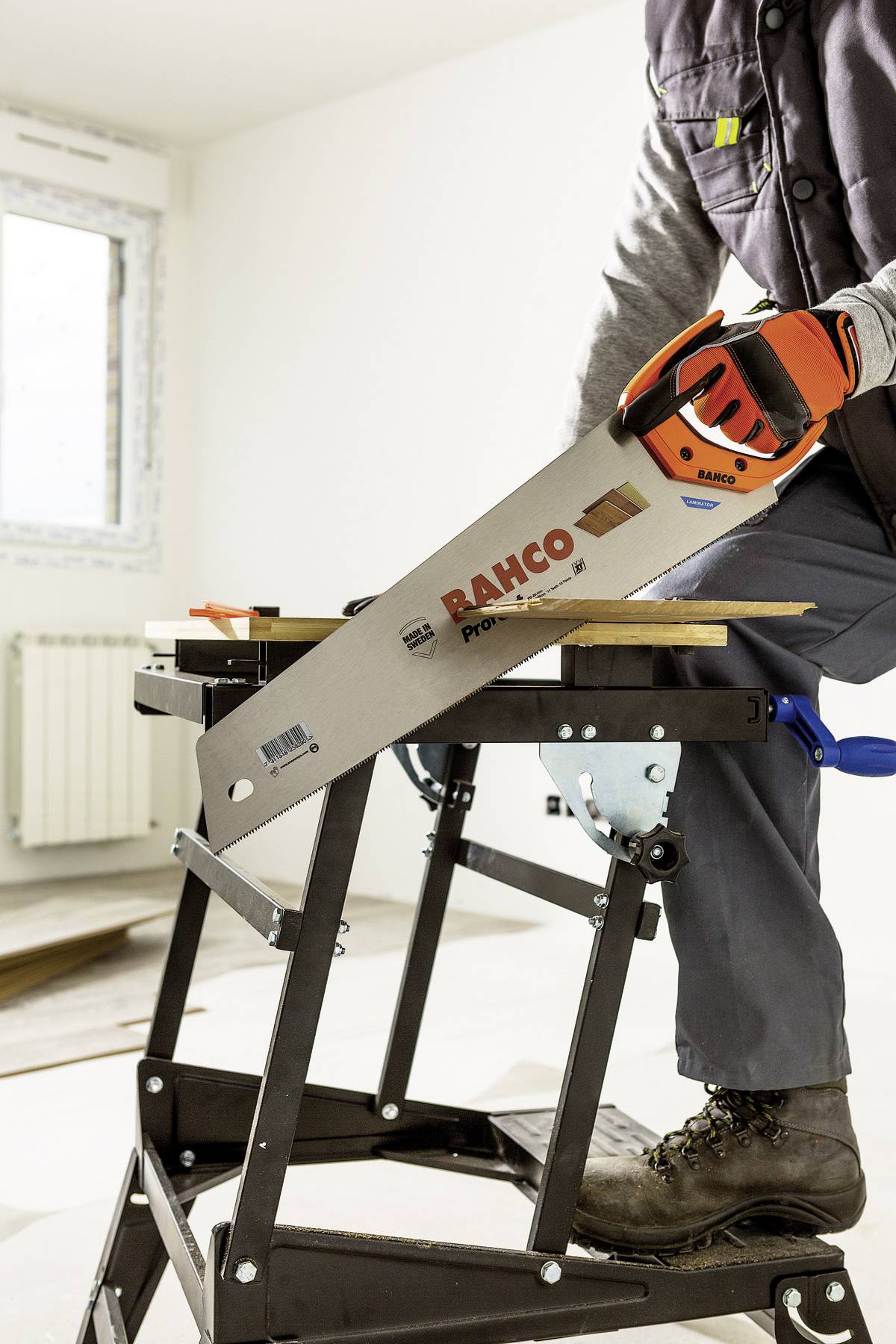 Eine Person sägt mit einer Handsäge ein Holzbrett auf einem Klapptisch in einem hellen Raum. Die Säge trägt die Aufschrift 'RAHCO'.