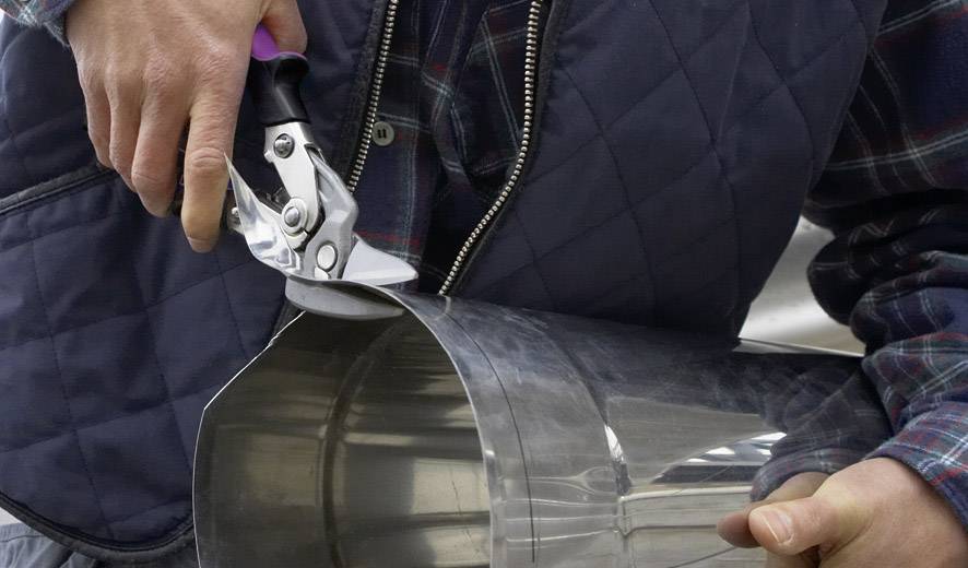 Une personne coupe une tôle argentée avec une cisaille à métaux. Elle porte un gilet matelassé et une chemise à carreaux.
