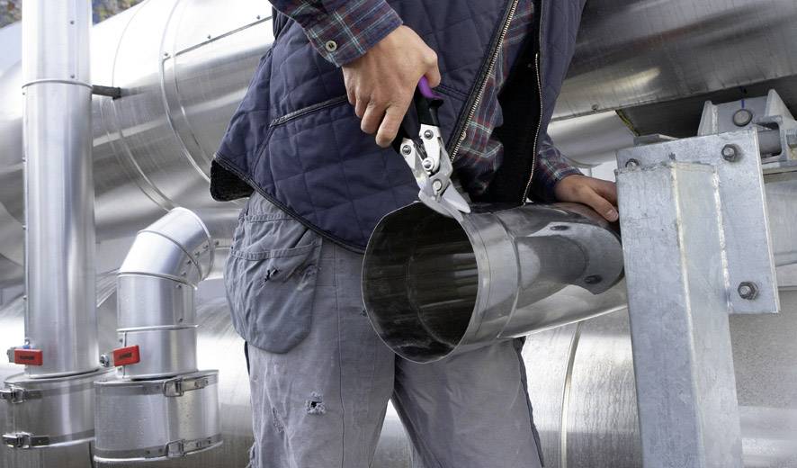Une personne coupe un tuyau métallique avec des cisailles à métaux sur un chantier de construction. À l'arrière-plan, d'autres tuyaux métalliques sont visibles.