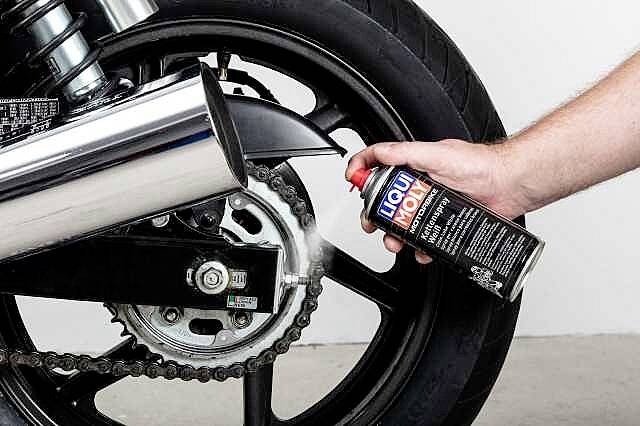 Une main vaporise du dégraissant pour chaîne à partir d'une bombe aérosol sur la chaîne d'une moto noire.