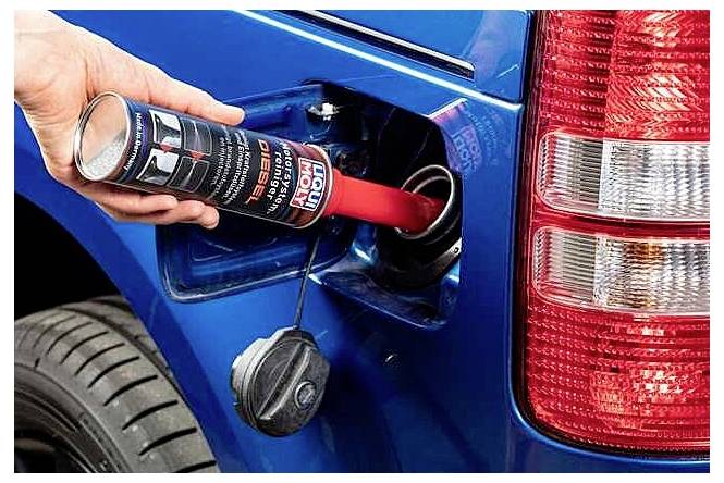 Une main verse un additif pour carburant diesel d'une bouteille dans l'ouverture du réservoir de carburant d'un véhicule bleu situé à côté du feu arrière.