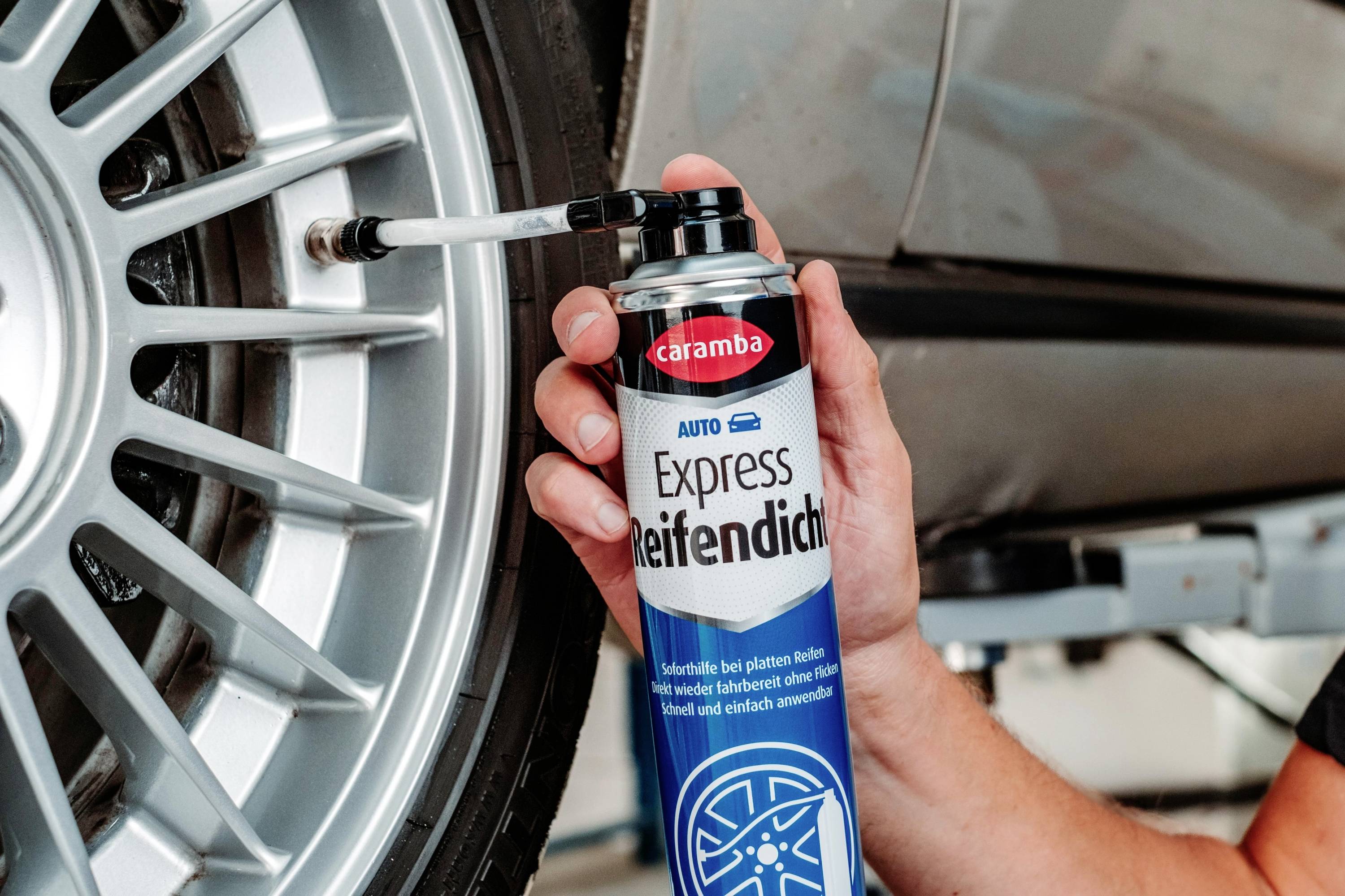Personne utilisant une bombe de 'Caramba Auto Express Reifendicht' pour réparer un pneu de voiture sur un pont élévateur dans un environnement de garage.