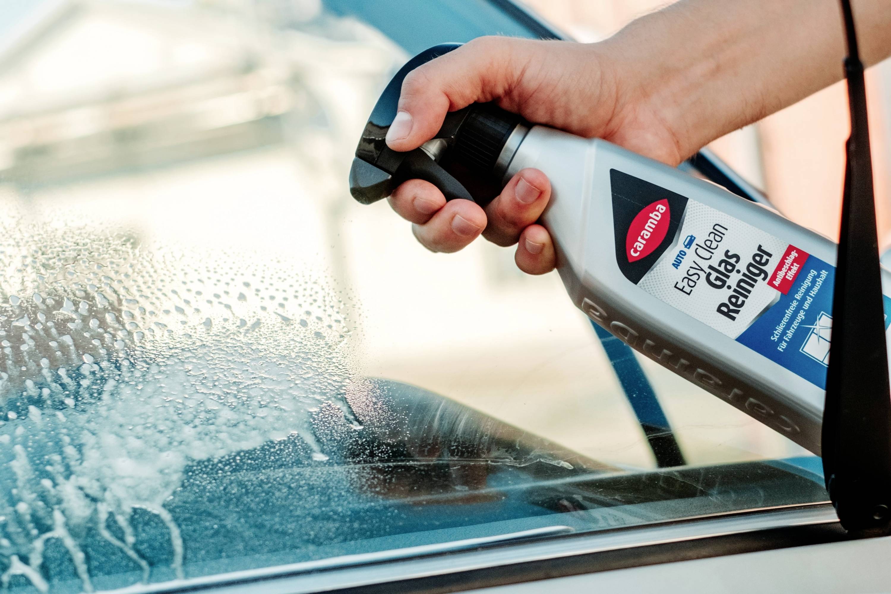 Une personne vaporise du nettoyant pour vitres sur un pare-brise de voiture, créant de la mousse. La bouteille de produit de nettoyage est étiquetée « Easy Clean Glas Reiniger ».