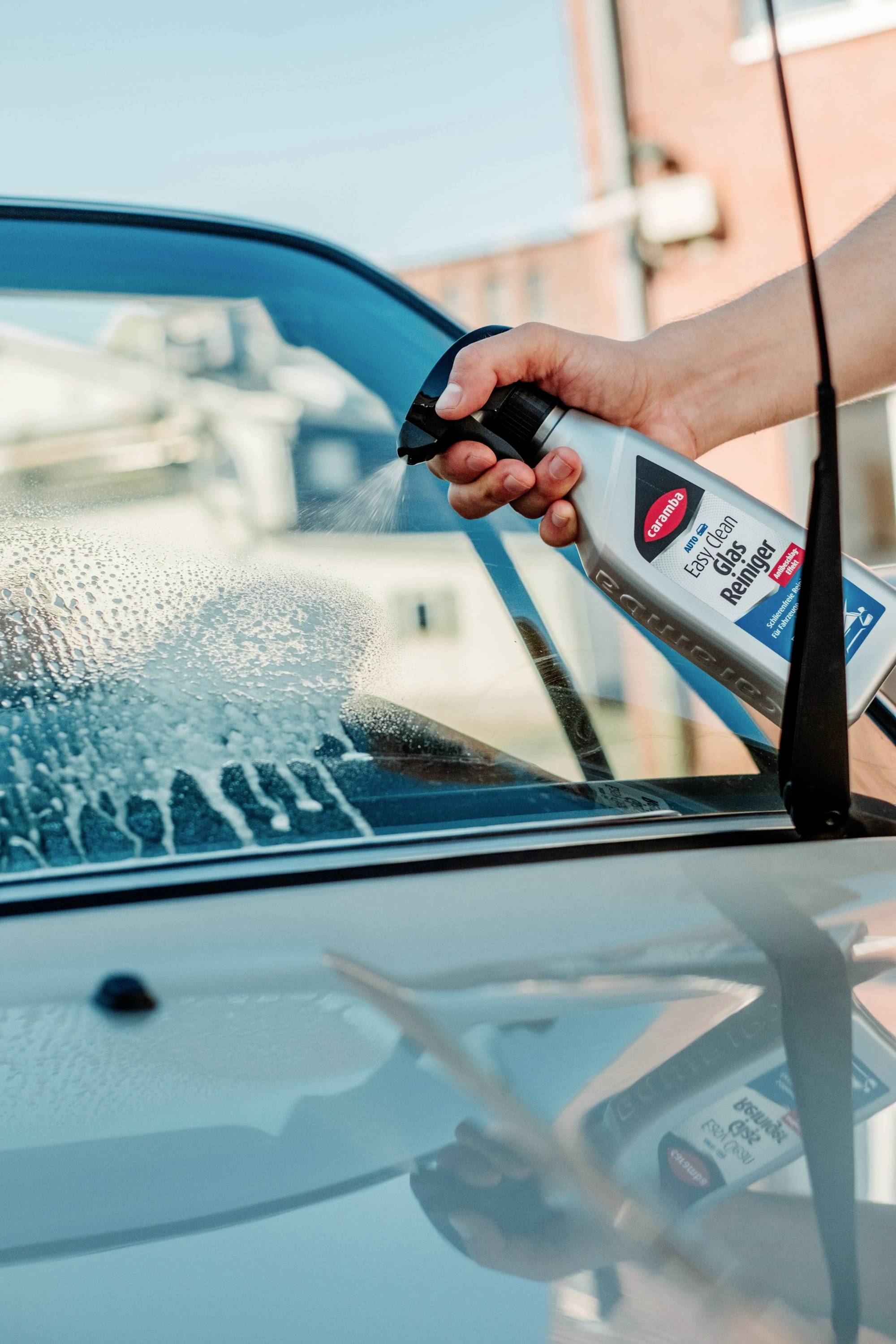 Une personne vaporise du produit nettoyant pour vitres sur un pare-brise de voiture, en préparation du nettoyage. Un bâtiment et le ciel sont visibles en arrière-plan.