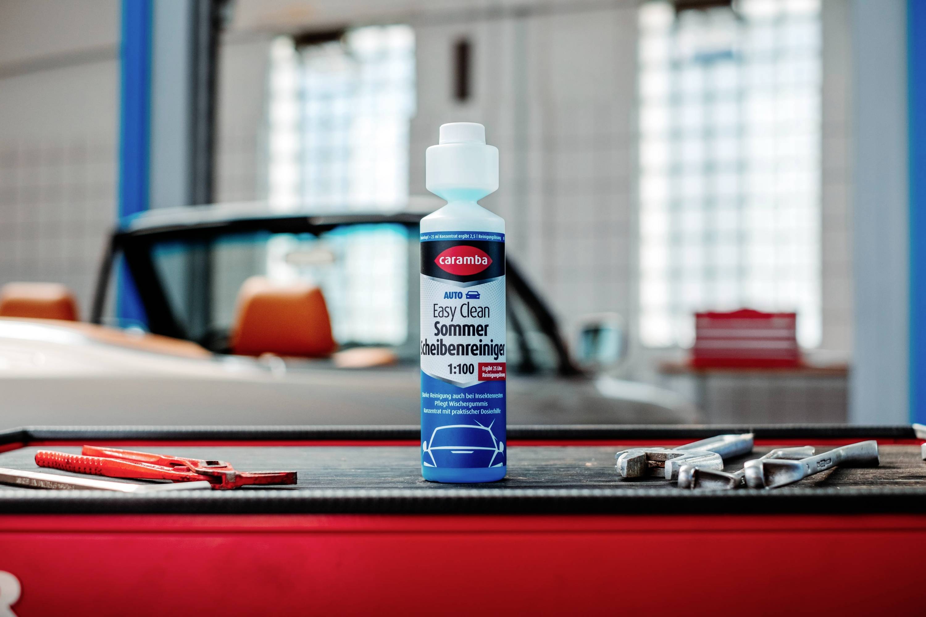 Une bouteille de liquide lave-glace 'Easy Clean Sommer Scheibenreiniger' posée sur un établi dans un garage, avec une voiture floue en arrière-plan.