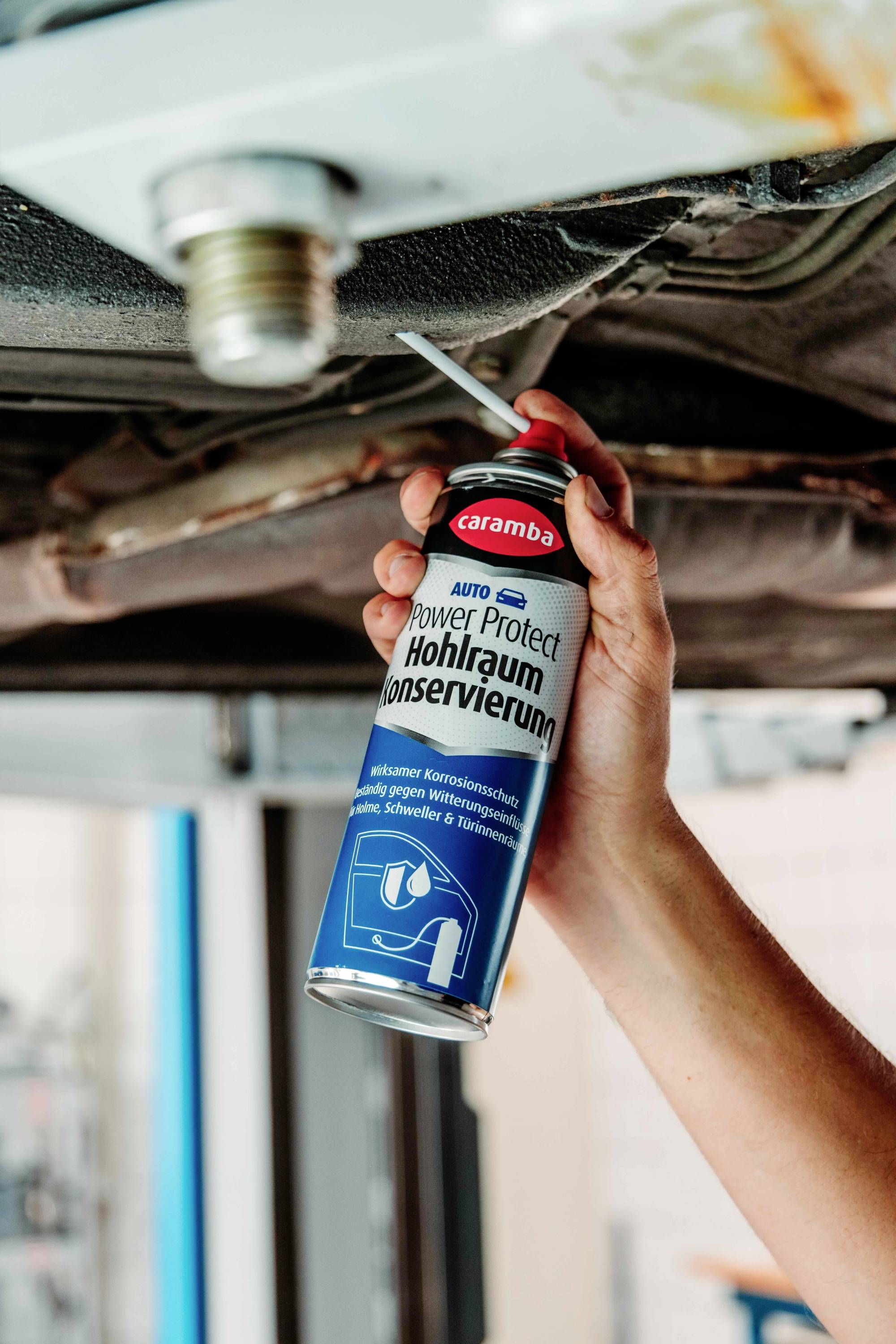 Une personne pulvérise un produit anticorrosion de préservation des cavités à partir d'une bombe aérosol sur le soubassement d'une voiture dans un garage, indiquant un travail de maintenance.