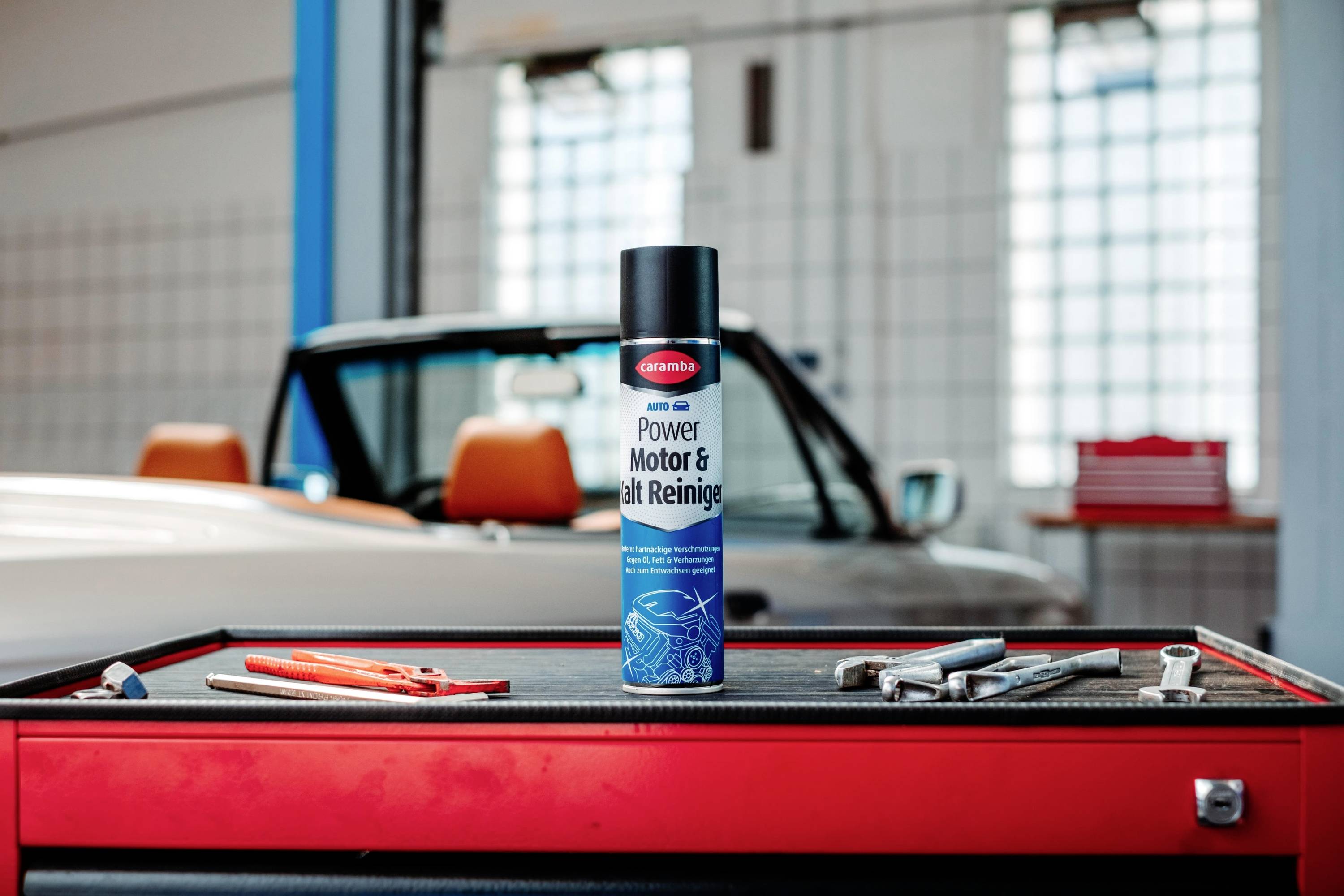Bombe aérosol étiquetée « Power Motor & Ent Reiniger » sur une boîte à outils d'atelier. Une voiture et des outils sont visibles en arrière-plan.