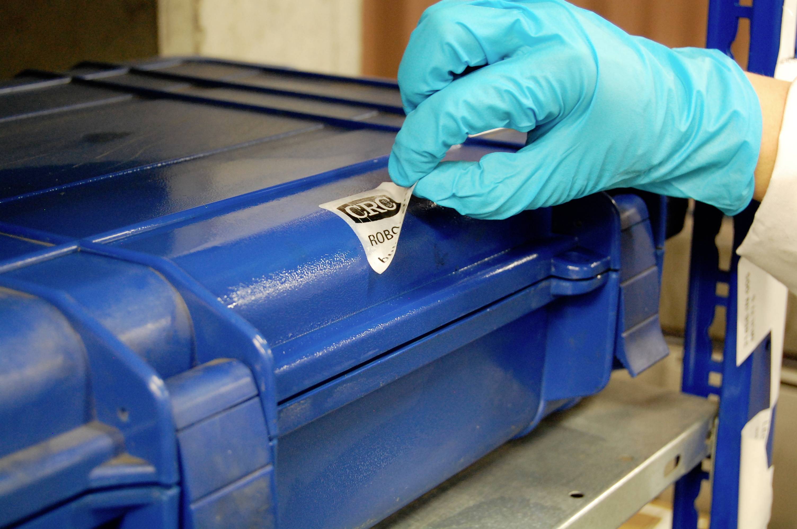 Une main gantée retire un sceau de sécurité d'une valise bleue fermée. La valise est posée sur une étagère.