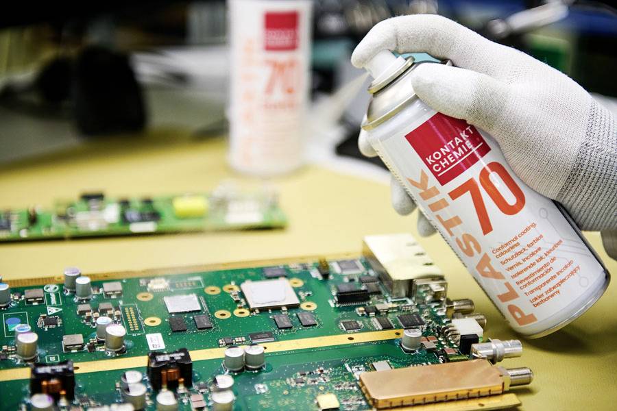 Une main gantée pulvérise du « Plastik 70 » sur une carte de circuit imprimé verte, suggérant une protection ou une maintenance des composants électroniques.