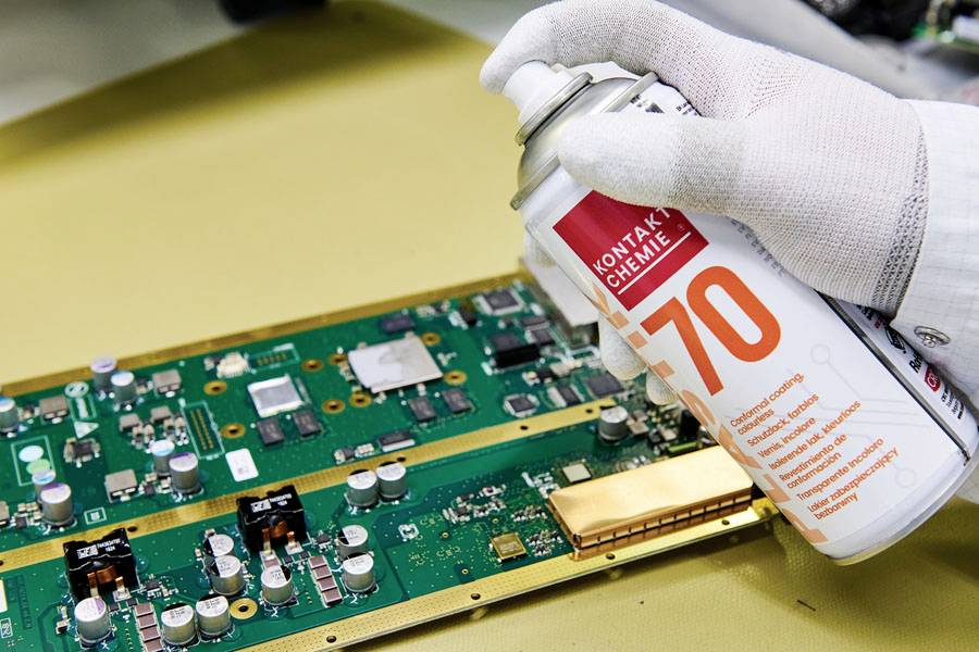 Une main gantée vaporise un nettoyant électronique sur une carte de circuit imprimé verte posée sur un établi, illustrant l'entretien de composants électroniques.