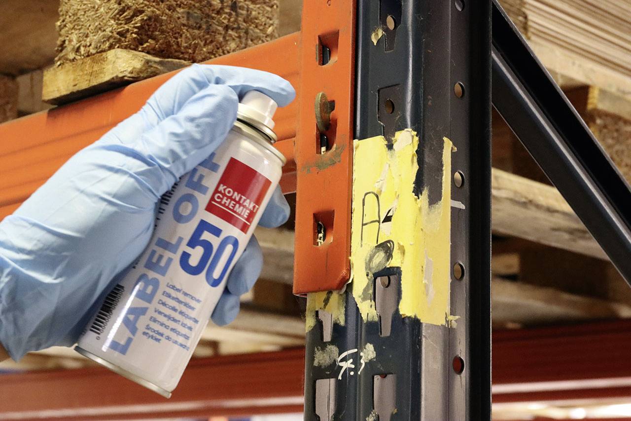 Une main gantée de bleu pulvérise un aérosol 'Label Off 50' sur une étagère métallique afin de retirer les résidus de colle.