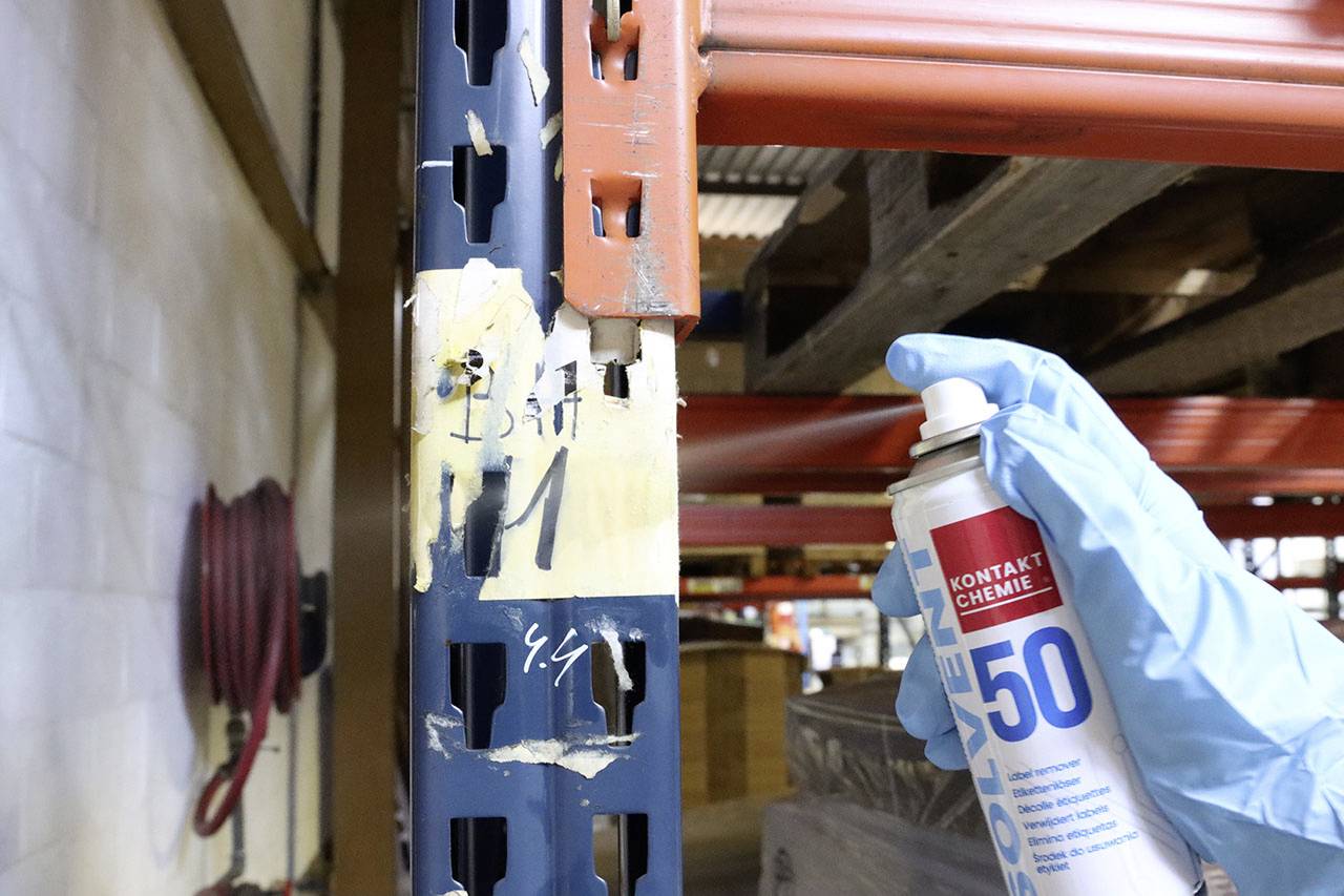 Une personne porte des gants bleus et tient une bombe de lubrifiant pour entretenir une étagère de rangement en métal.