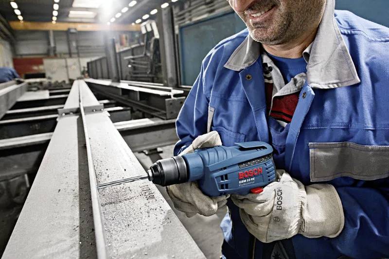 Une personne dans un atelier, souriant tout en utilisant une perceuse électrique bleue Bosch sur une poutre métallique, portant des gants de protection et une veste bleue.