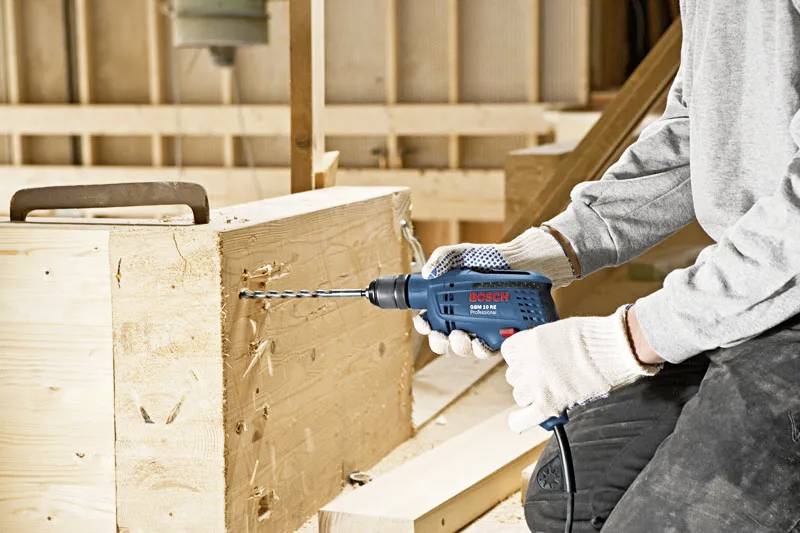 Une personne portant des gants perce un trou dans une poutre en bois à l'aide d'une perceuse électrique dans un environnement d'atelier.