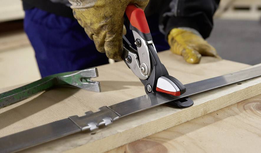 Une personne coupe une bande d'acier avec des ciseaux métalliques sur une planche en bois. Elle porte des gants jaunes comme mesure de protection.