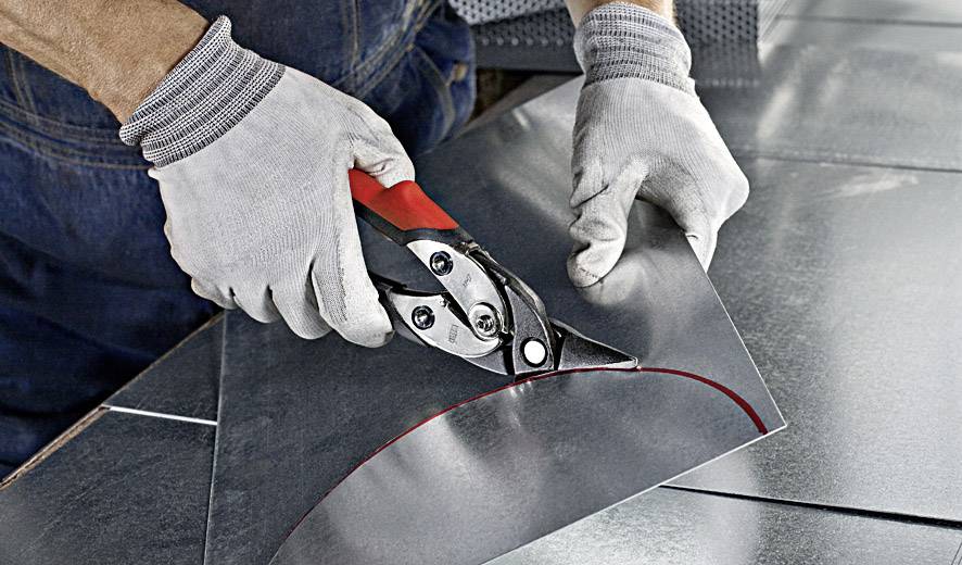 Une personne coupe un morceau de tôle avec des cisailles métalliques. Les mains portent des gants de protection afin d'éviter les blessures.