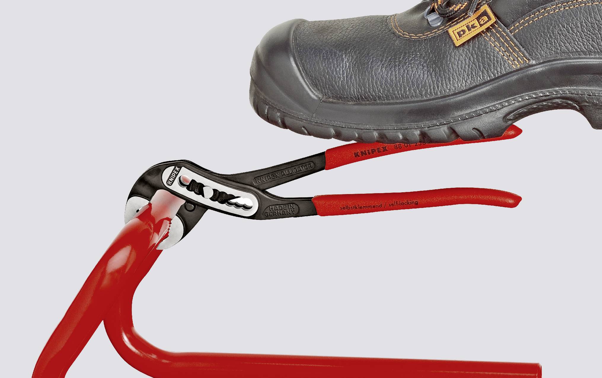 Une chaussure de sécurité noire maintient un tuyau rouge à l'aide d'une pince rouge.