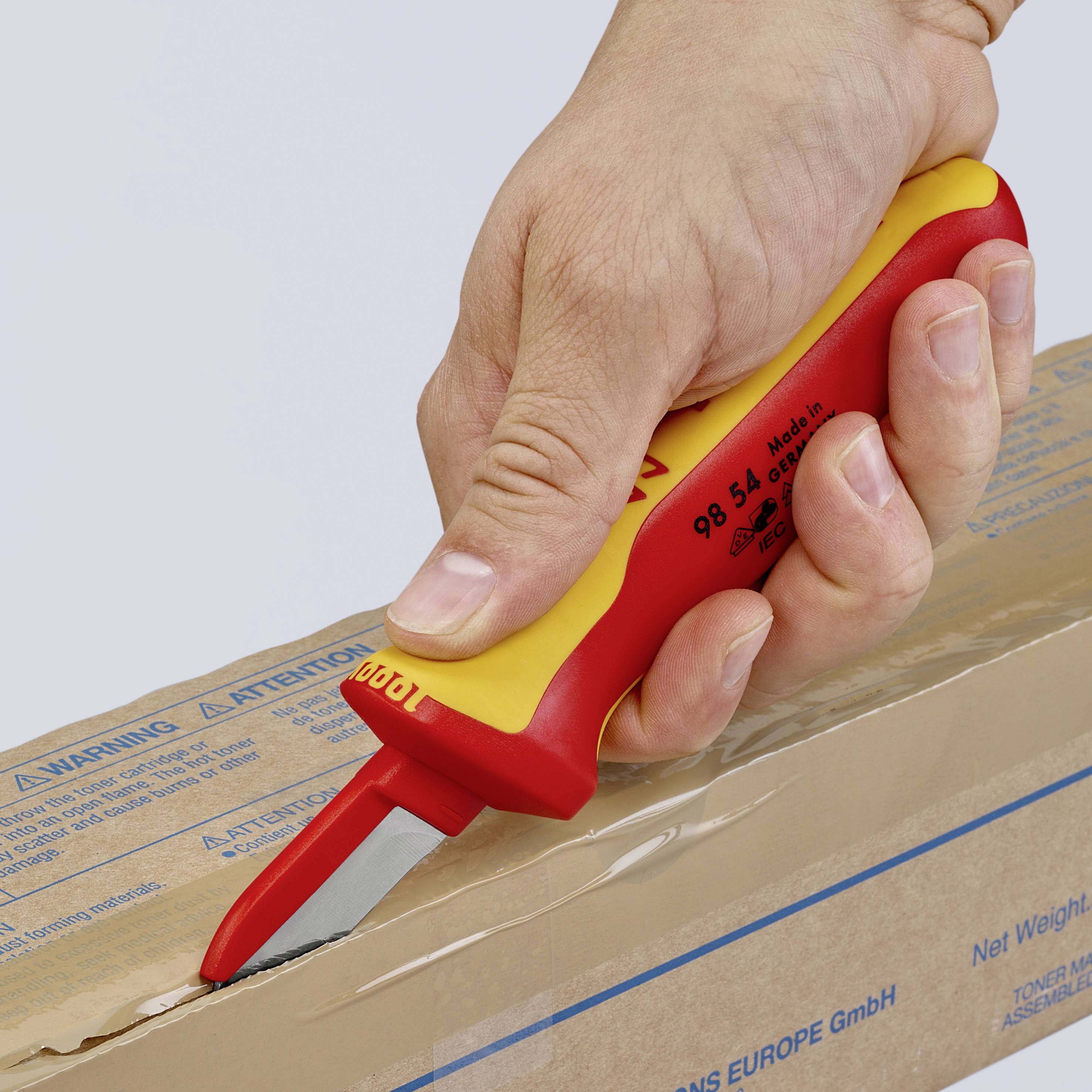 Une main coupe un carton avec un couteau de sécurité jaune et rouge. « Avertissement : changement de lame » est inscrit sur le couteau.