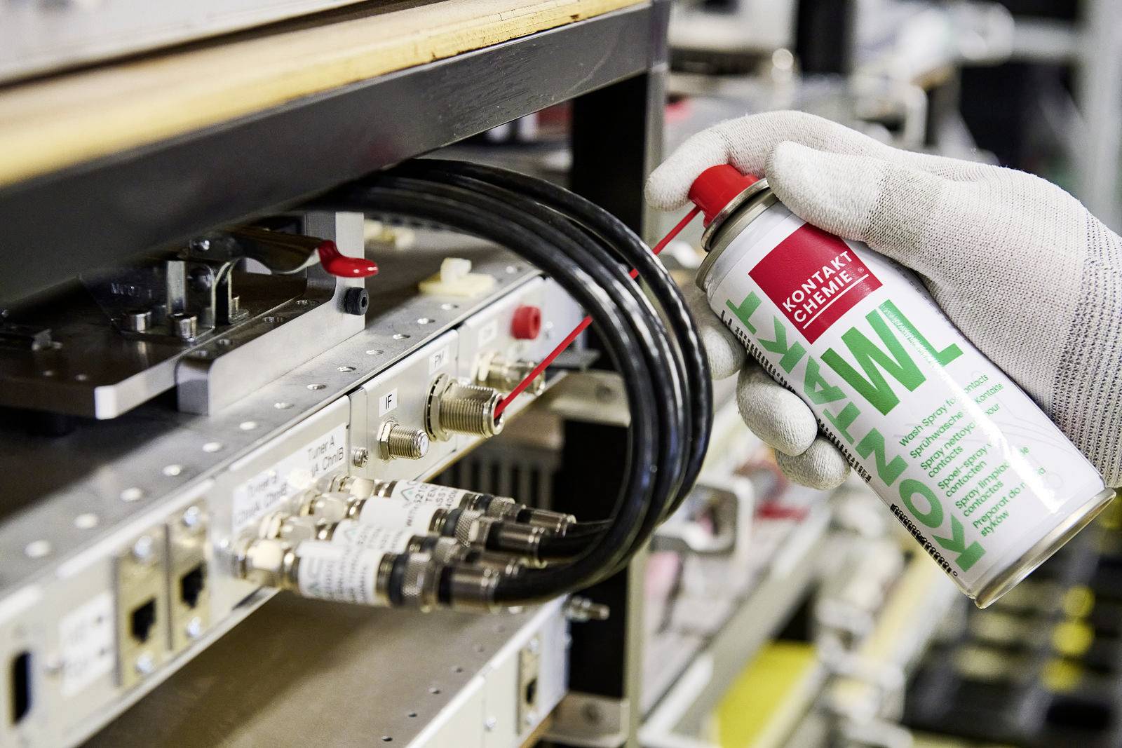 Une main gantée vaporise un spray de contact sur des connexions électroniques dans un dispositif technique afin d'améliorer la conductivité.