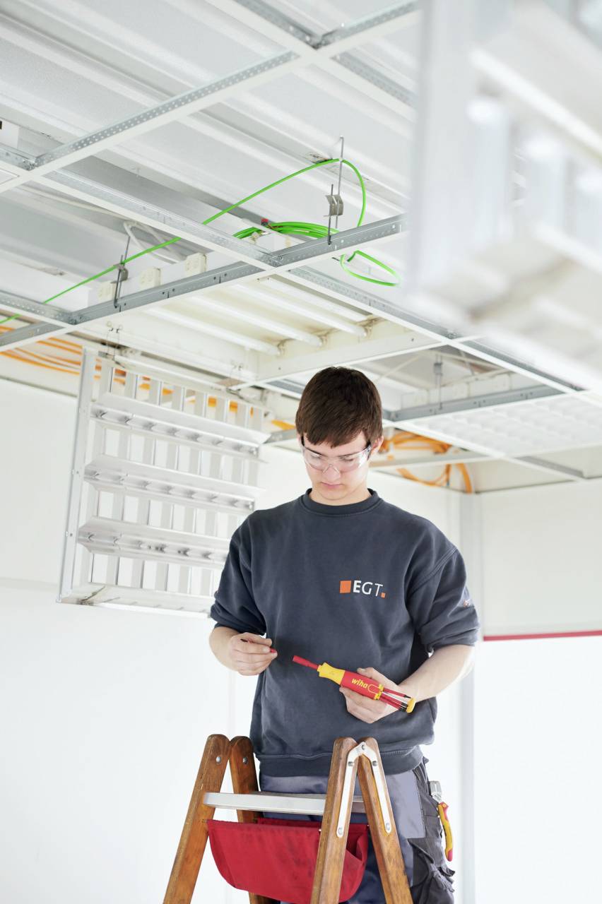 Un électricien est debout sur une échelle et travaille sur le câblage du plafond dans un espace industriel.