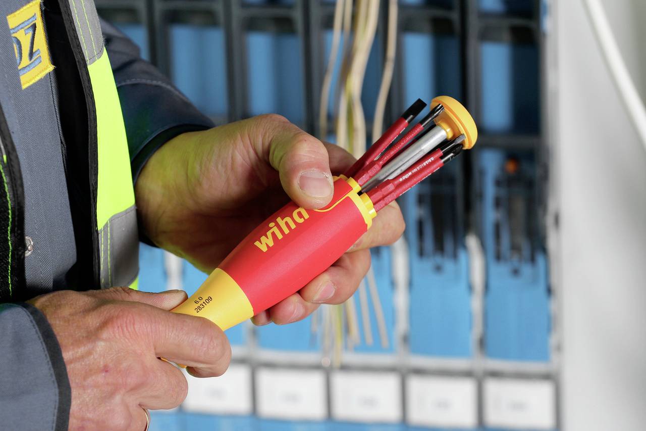 Une personne tient un tournevis rouge Wiha avec de nombreux embouts interchangeables dans une main devant un coffret électrique.