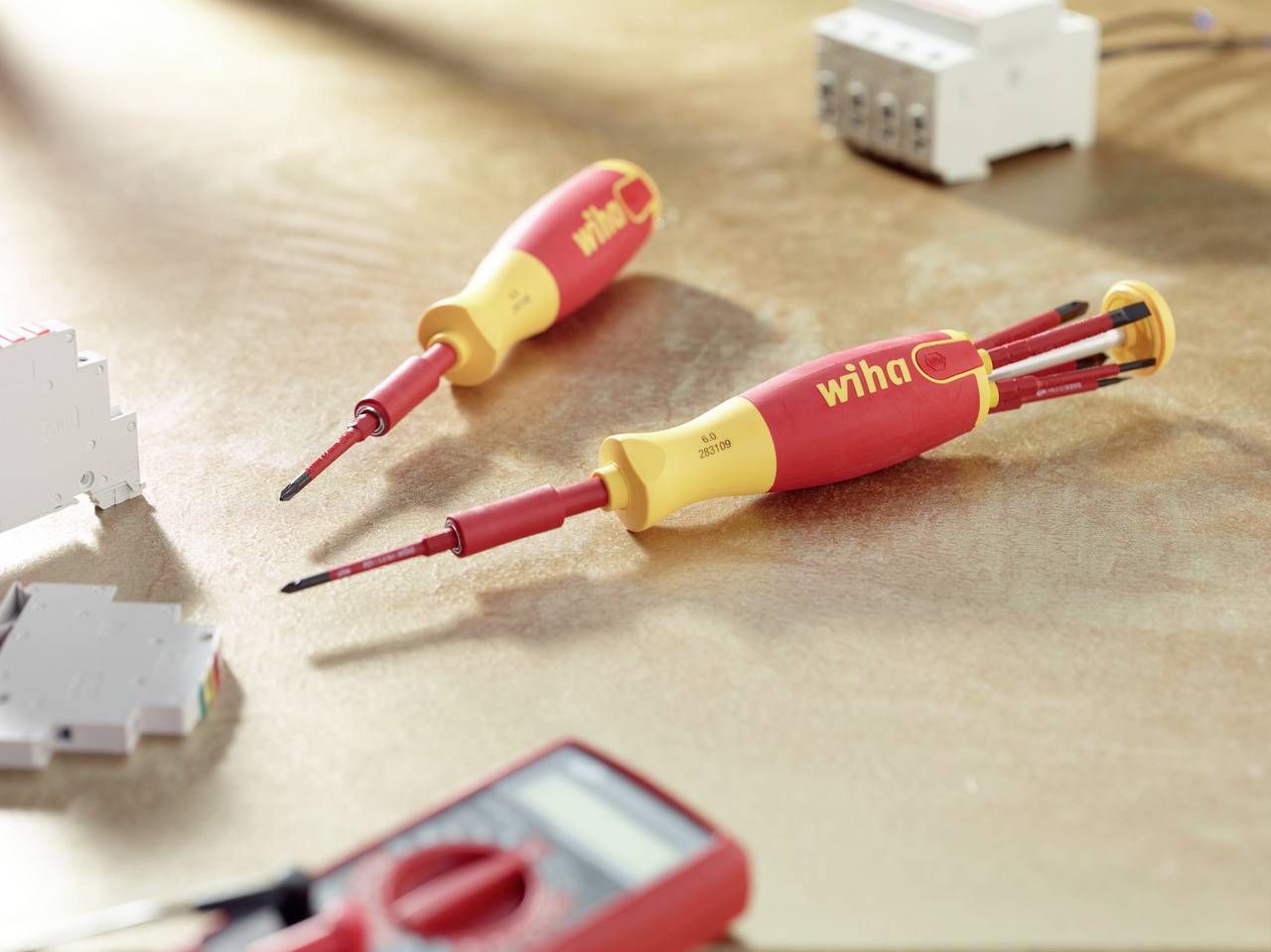 Deux tournevis rouges avec des poignées jaunes sont posés sur une table, à côté d'un appareil de mesure électrique. Des accessoires techniques sont visibles en arrière-plan.